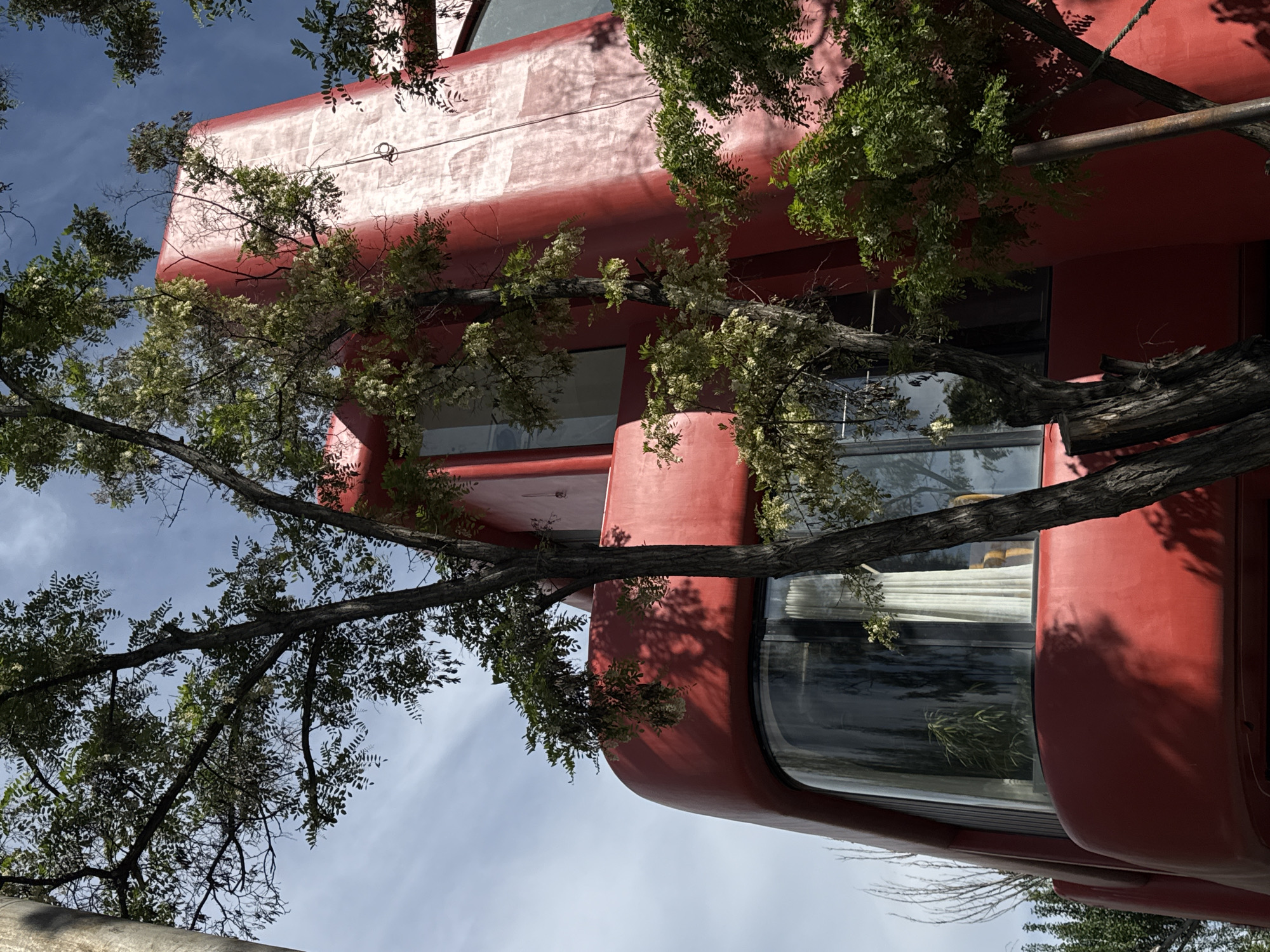 A low-angle shot depicts a red, multi-story building situated in Yerevan, Armenia. A leafless tree branch extends diagonally across the frame, partially obscuring the building. The structure exhibits rounded corners and smooth surfaces, with large windows visible on at least two levels. The sky, light blue with wispy clouds, is visible behind the building and through the tree branches. A metal cable runs along the upper portion of the building.