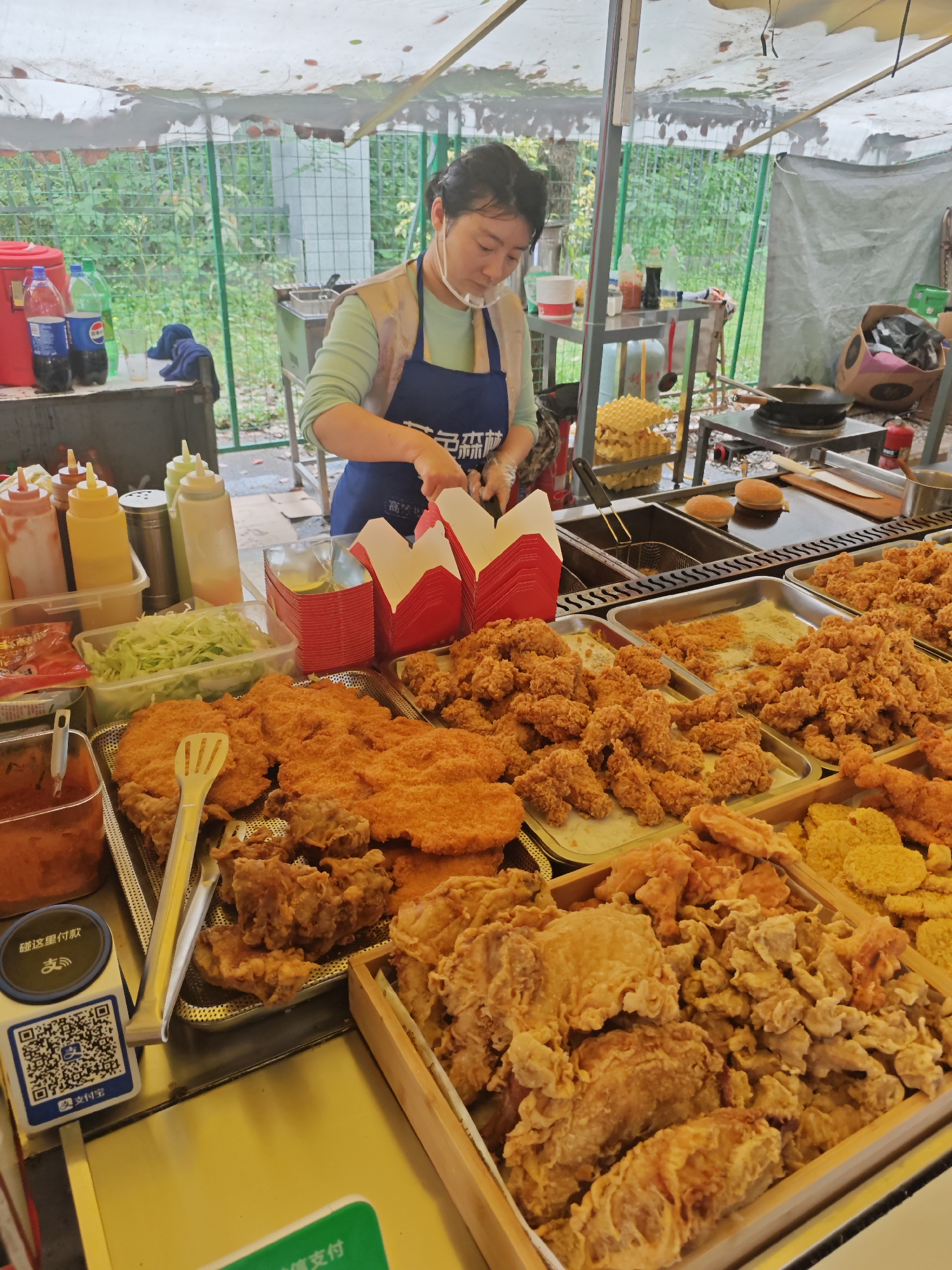 The image depicts a street food vendor in the Shapingba District of China. An Asian woman wearing a face mask and blue apron is packing food into red containers. In front of her is an array of fried foods: chicken, cutlets, and other items in metal trays and a wooden box. Various condiments, including sauces and shredded lettuce, are arranged nearby. The vendor operates under a canopy. A payment kiosk is on the left. Cooking equipment, including a deep fryer and a flat-top grill, is visible in the background. A fence with green foliage is further in the background.