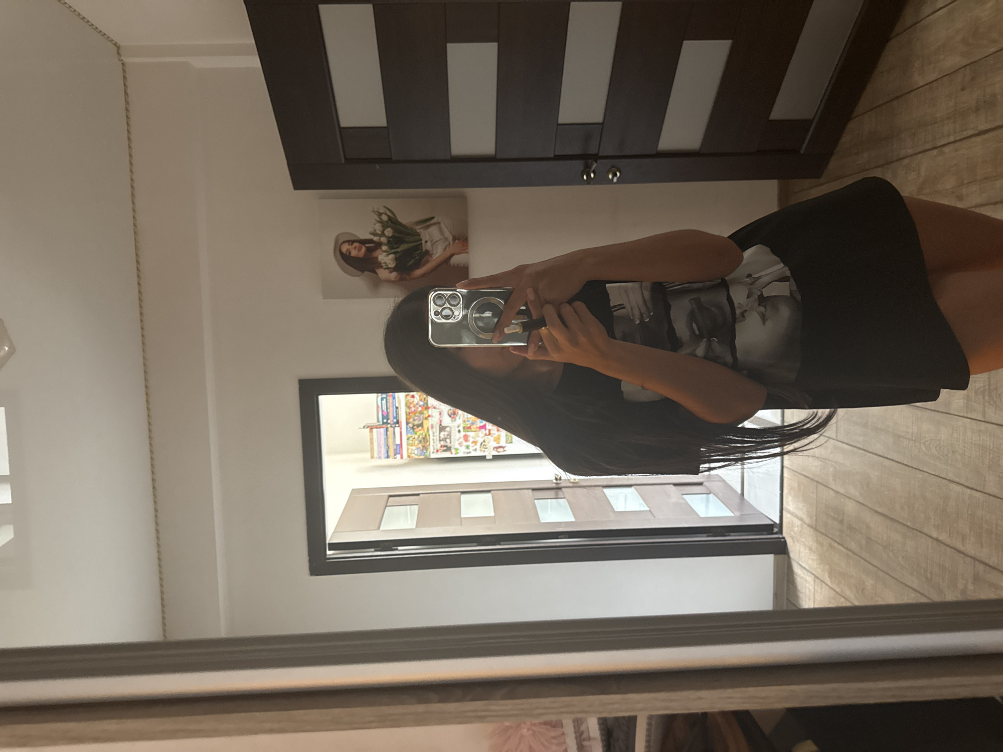 In an indoor scene, a young woman with long, dark hair takes a mirror selfie. She is wearing a black t-shirt with a graphic print and a black skirt. She is holding a smartphone in her right hand, with her left hand pointing to the screen. Behind her, a dark brown door with glass panes is visible on the right. To the left of the door, a framed picture of a woman holding tulips is hanging on the wall. Further to the left, another door is ajar, revealing a room with shelves filled with colorful items. The floor is made of light-colored wooden planks. The overall environment suggests a residential interior. There is no discernible information in the image that would place this scene in Chișinău, Moldova.