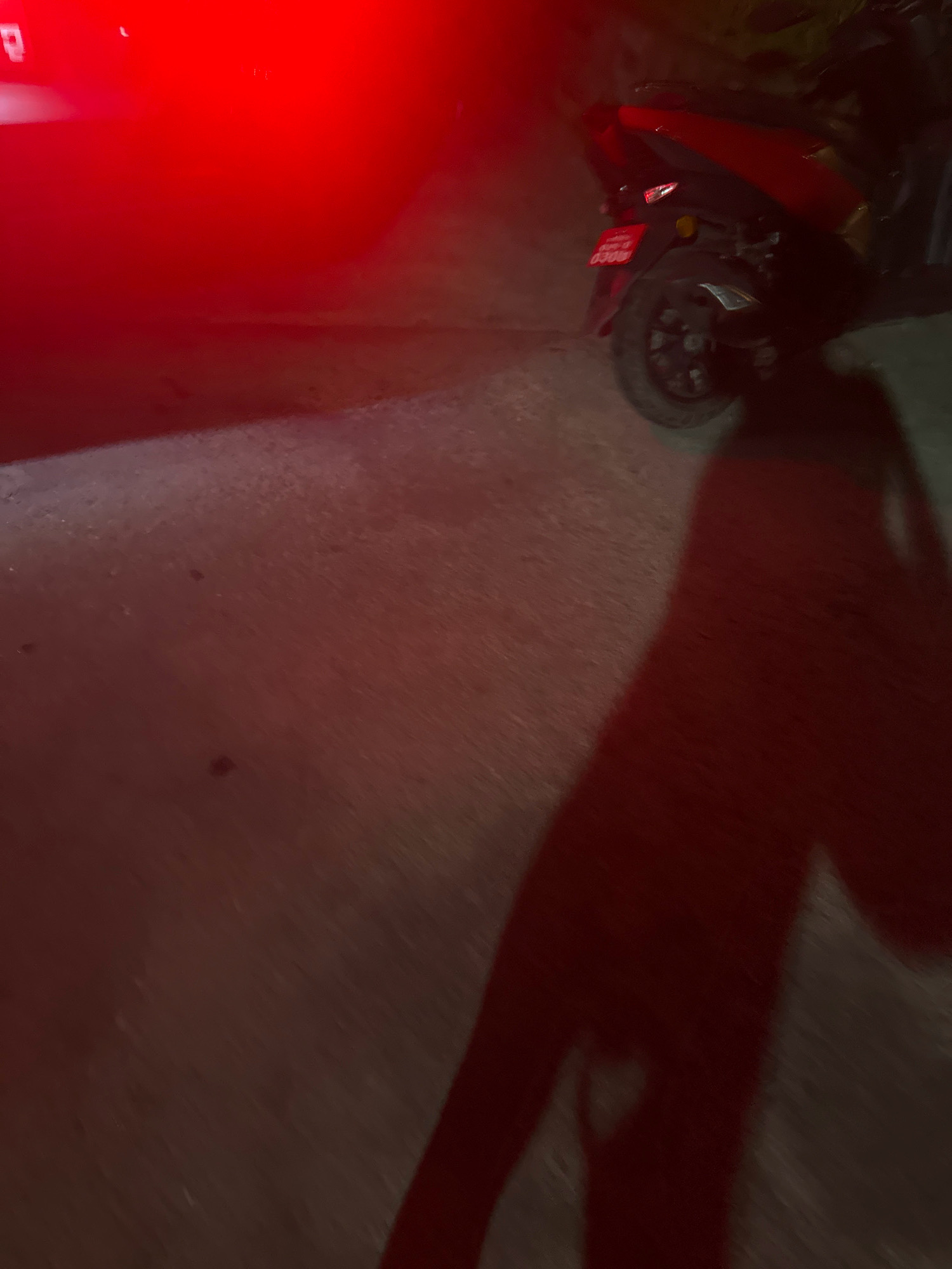 The image shows a red scooter parked on a paved surface at night. A bright red light, likely from the scooter's taillight or a nearby source, illuminates the scene. A dark shadow of a person is cast across the ground and the side of the scooter. The license plate on the scooter is visible, though the numbers are not clearly legible. The background is dark, with some vegetation visible on the right side. The image appears to have been taken in Lalitpur, Nepal, based on the style of the license plate.