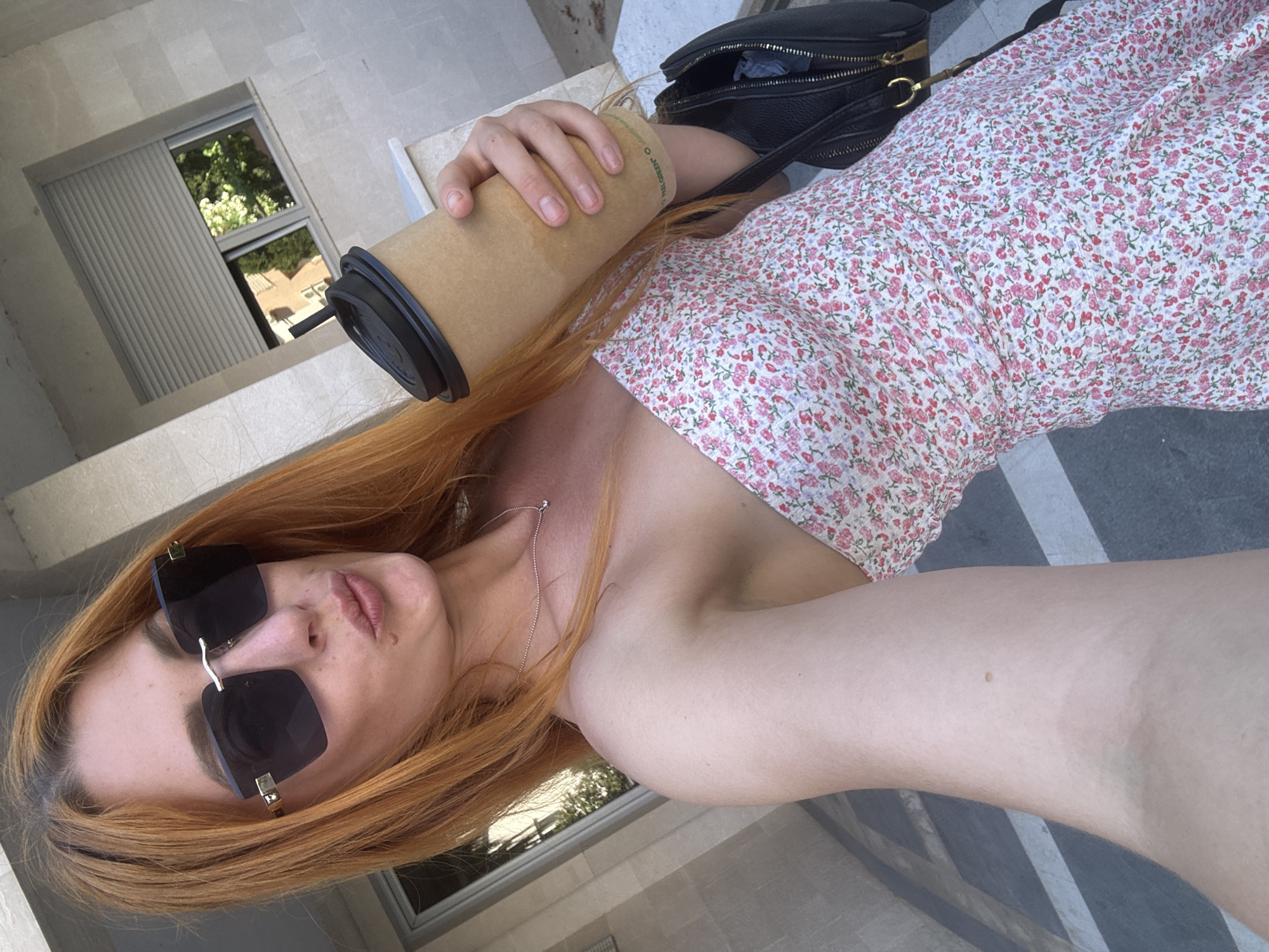 A young woman with auburn hair and sunglasses is holding a coffee cup and taking a selfie outdoors. She is wearing a strapless floral dress and a necklace. A black bag is visible over her shoulder. The background suggests an urban environment, with architectural details and a window reflecting trees. The image appears to be taken during the day.
