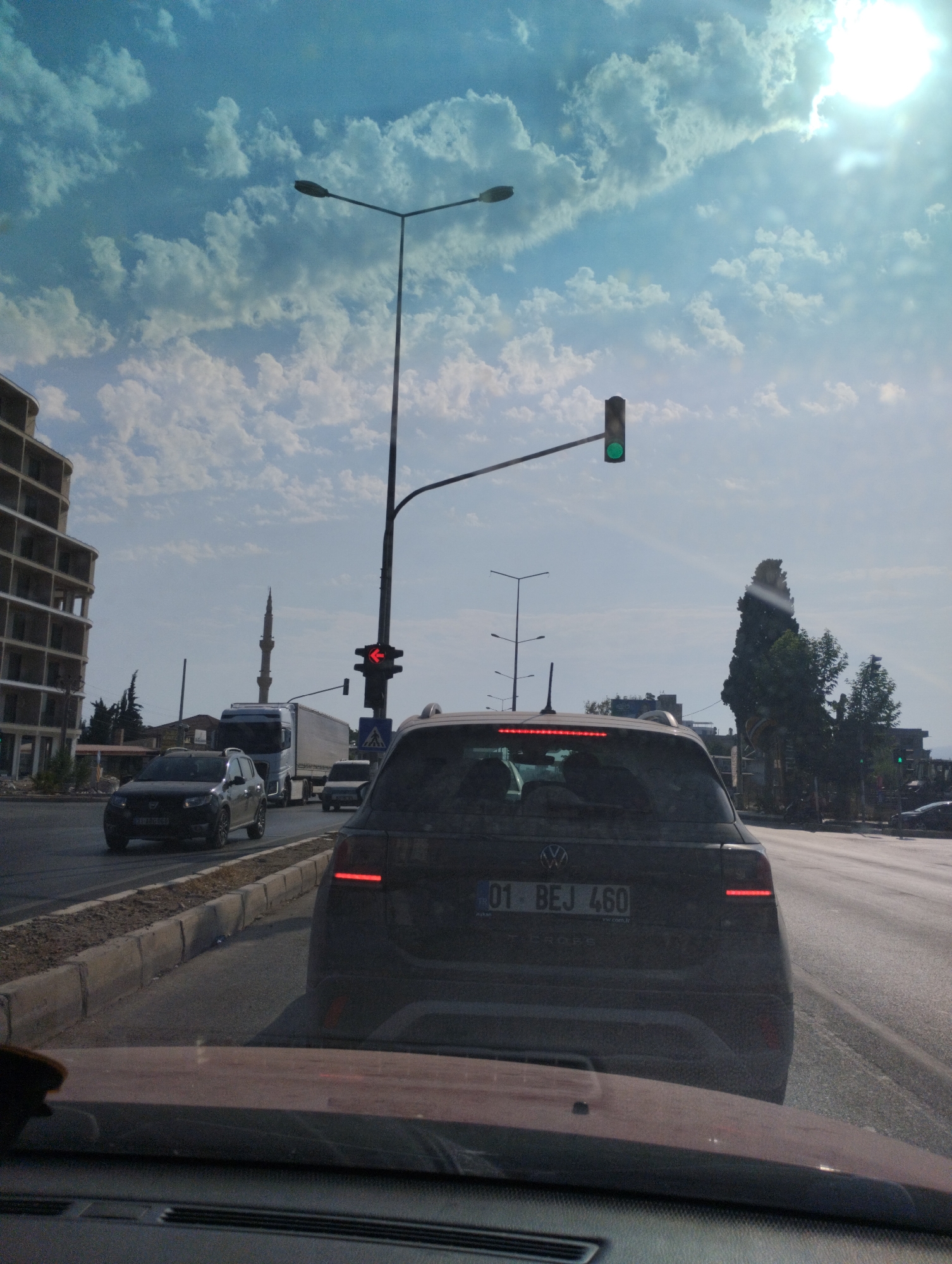 The image is taken from the perspective of a driver in Erdemli, Turkey. The foreground shows the dashboard of a car, with a blurred reddish-brown surface. In the middle ground, the rear of a dark-colored Volkswagen T-Cross is visible, with its license plate reading "01 BEJ 460". The car's brake lights are illuminated. Traffic is present on a paved road, with other vehicles including a dark SUV and a white truck. To the left, a multi-story building under construction with exposed concrete floors and columns is visible. A tall, slender minaret of a mosque can be seen behind the construction. Streetlights and traffic signals are also present. The sky is a vibrant blue with scattered white clouds, and the sun is shining brightly, creating some lens flare. The environment appears to be an urban setting with a clear sky.