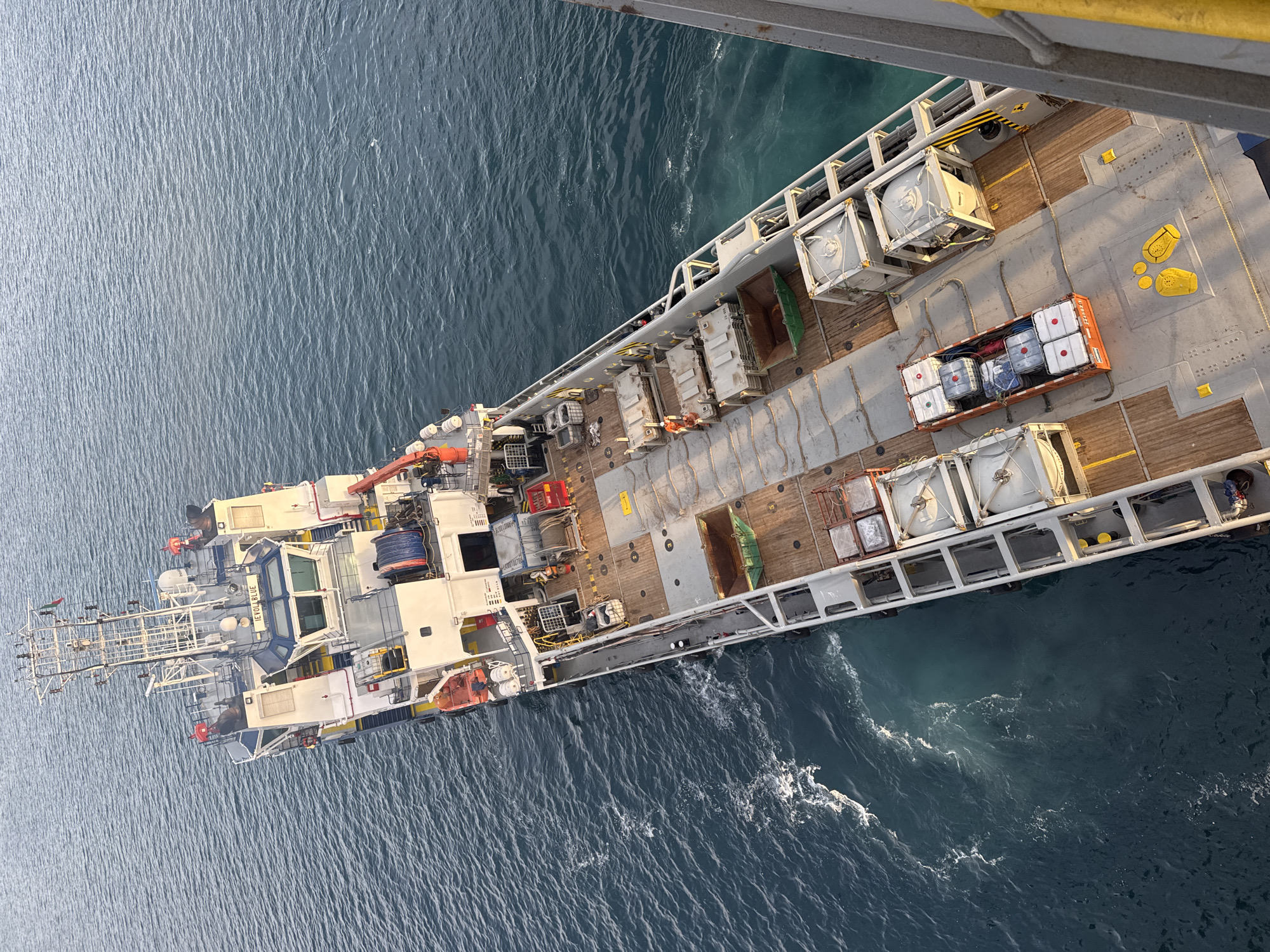 This is a top-down, slightly angled aerial view of a large vessel, possibly a supply ship, sailing on a dark blue, choppy ocean. The vessel is oriented diagonally across the frame, with its bow pointing towards the upper left.
The deck of the ship is visible, featuring various equipment and cargo. Several large, rectangular white containers, some stacked, are secured to the deck. A red container filled with white cylindrical objects is also present. Yellow markings, including painted circles and striped areas, are visible on the deck. There are also some orange life rafts and other pieces of equipment, including cranes and structures that suggest a working vessel. The water is a deep blue with whitecaps, indicating moderate sea conditions.
The lighting suggests it might be late afternoon or early morning, with sunlight casting shadows on the deck. No people are clearly discernible. There is some text visible on the side of the ship, reading "IEVOY BLUE" and also smaller text on a container.