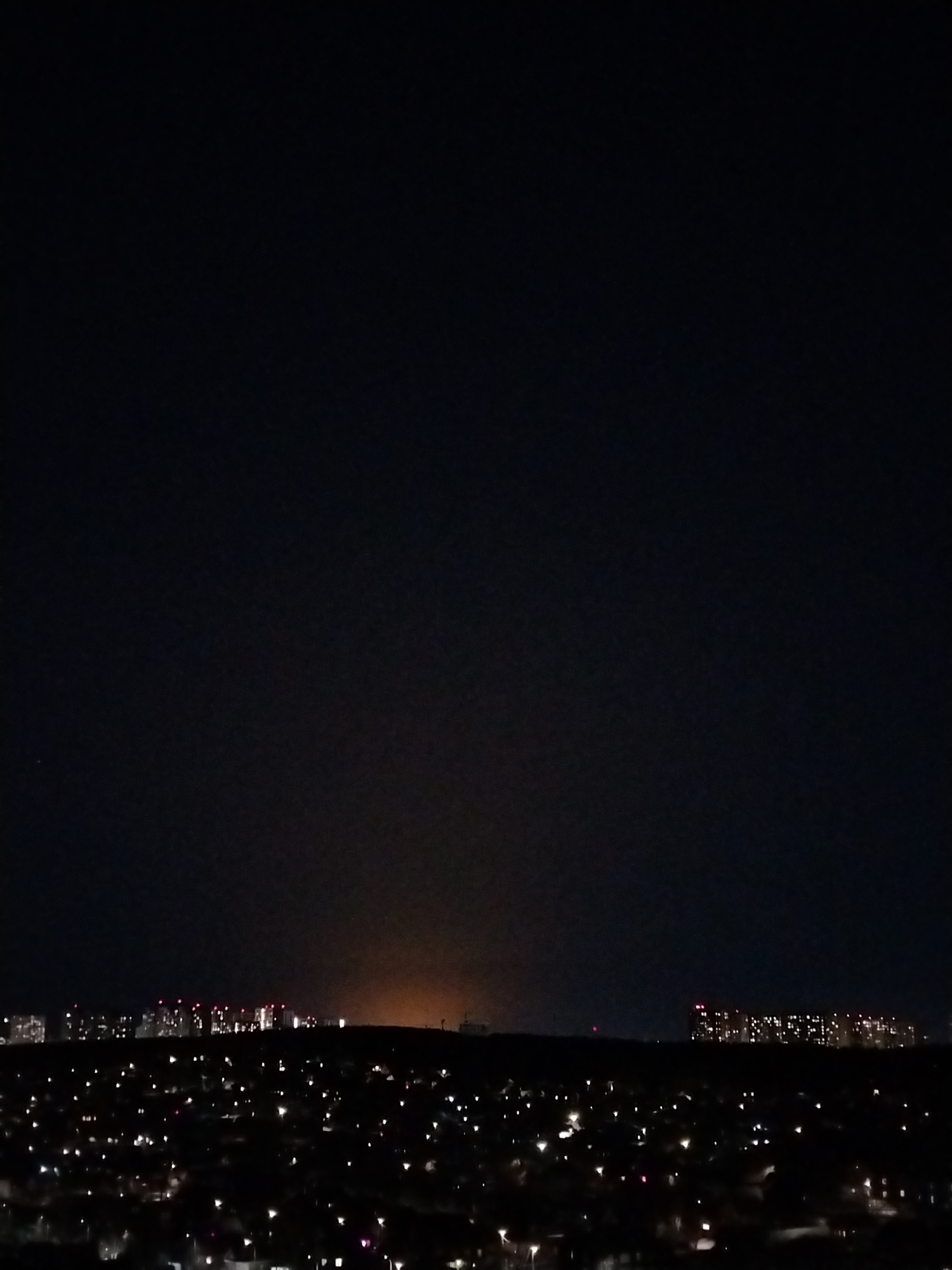 A night shot from a high vantage point overlooking a city. In the foreground, individual lights from houses and streets illuminate a residential area, contrasting with the dark landscape. In the distance, groups of taller buildings are visible, identifiable by their windows and illuminated rooftops. These buildings appear as flat, brighter shapes along the horizon. The skyline transitions into a dark, seemingly cloudless, night sky. The location is Izhevsk, Russia.