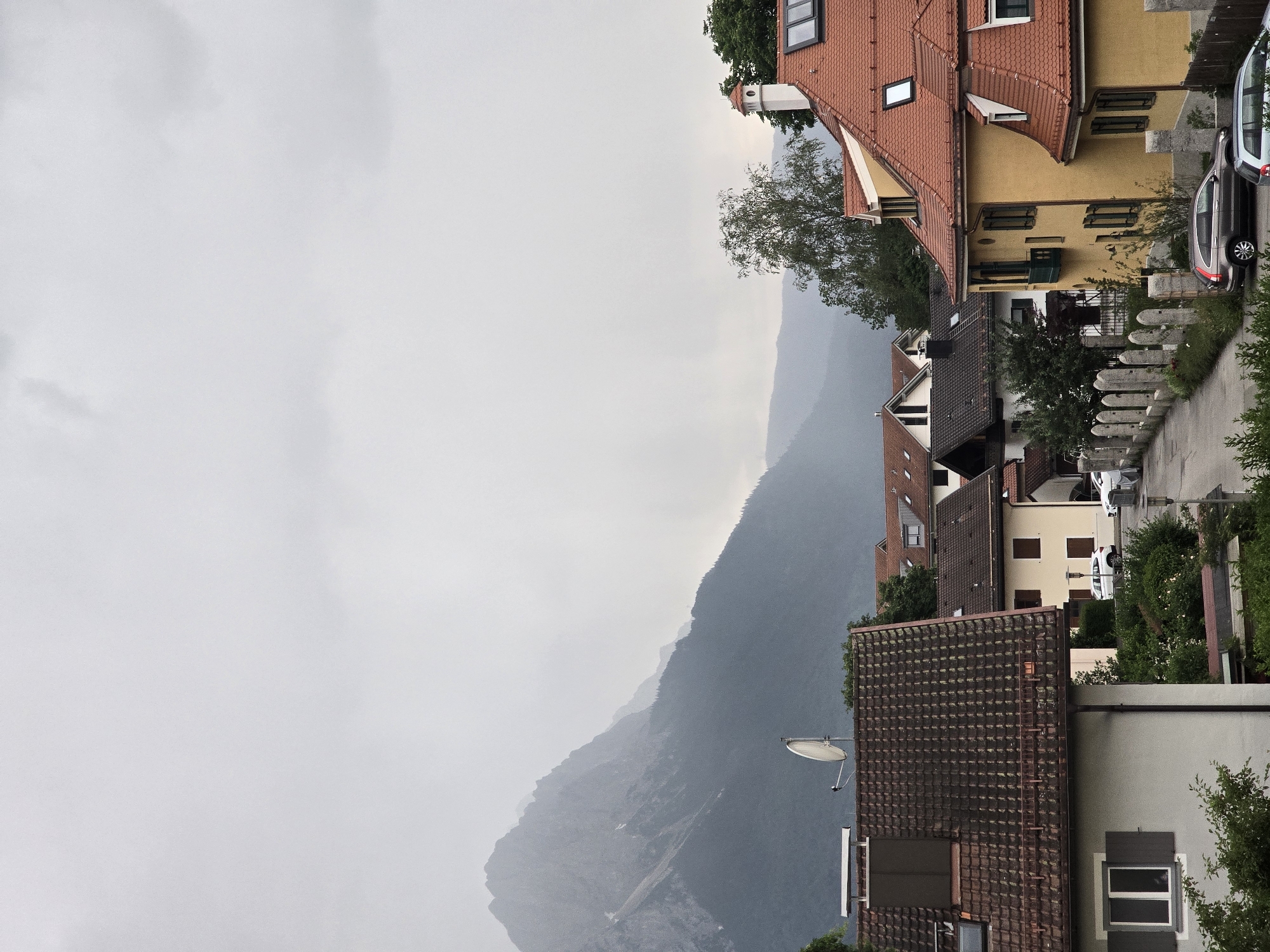 The image shows a view of a residential area with houses and mountains in the background. The houses have red tiled roofs and are of various architectural styles. Some of the houses have satellite dishes on their roofs. In the foreground, there are trees and a street with cars parked. The mountains in the background are covered in mist, suggesting a cloudy or rainy day. The scene is likely set in Garmisch-Partenkirchen, Germany, a town known for its mountainous landscape and traditional Bavarian architecture.