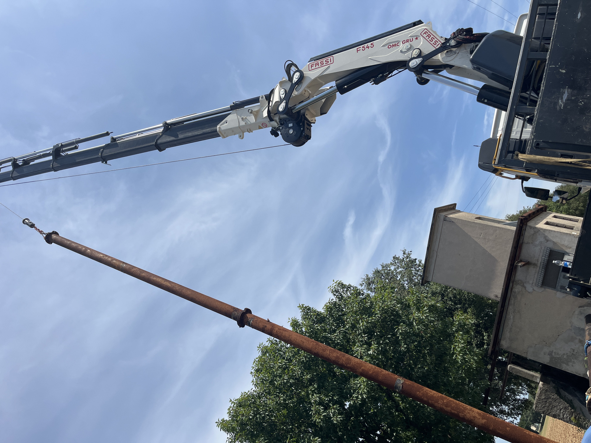 In the city of Castiglione delle Stiviere, Italy, under a partly cloudy blue sky, a long, rusty metal pipe is being lifted vertically by a crane. The crane, a Fassi F545 OMC GRU, is attached to the back of a truck. The pipe, approximately 10-15 meters long and 30 centimeters in diameter, is suspended from a hook at the end of the crane's boom. The pipe appears to be made of steel and shows signs of corrosion. In the background, there are some buildings and trees. The buildings are low-rise and appear to be industrial or utility structures. The trees are green and leafy. The overall scene suggests construction or demolition work is taking place.