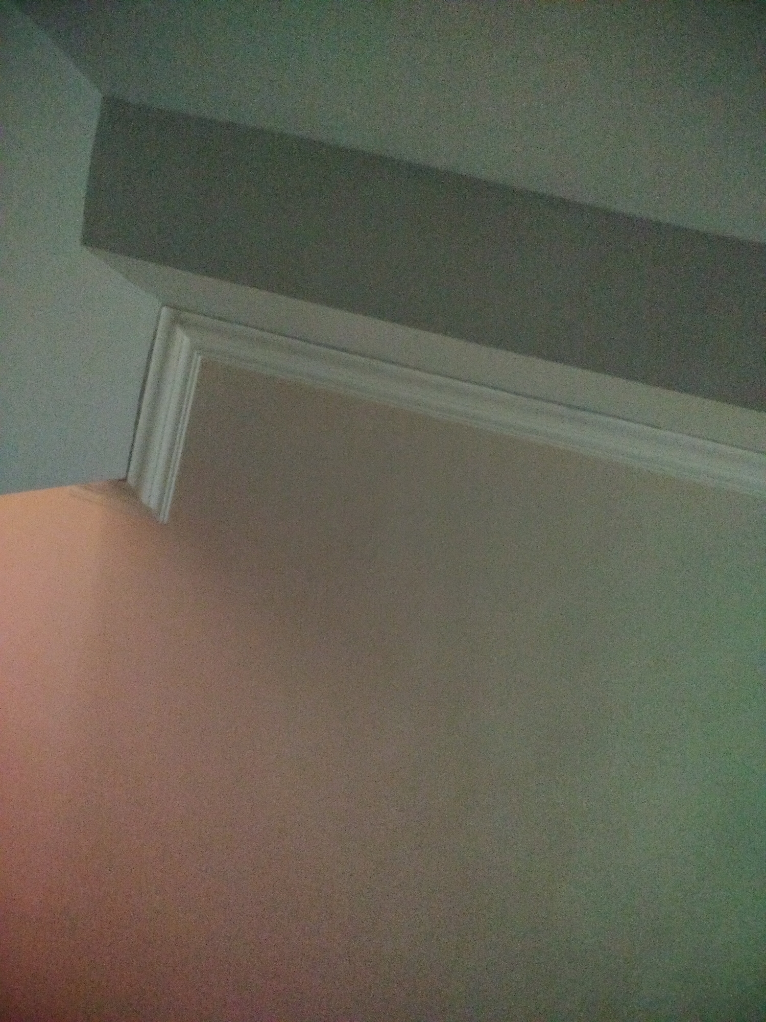The image shows a corner where a wall meets the ceiling. The wall is a light, neutral color, with a hint of pink in the lower left corner, possibly due to lighting. The ceiling is a lighter shade of gray. A white, decorative crown molding runs along the junction of the wall and ceiling. Above the molding, there's a horizontal structure, possibly a soffit, that projects from the ceiling. The image is taken from a low angle, looking up at the corner. There is no evidence of the location in Yerevan, Armenia.