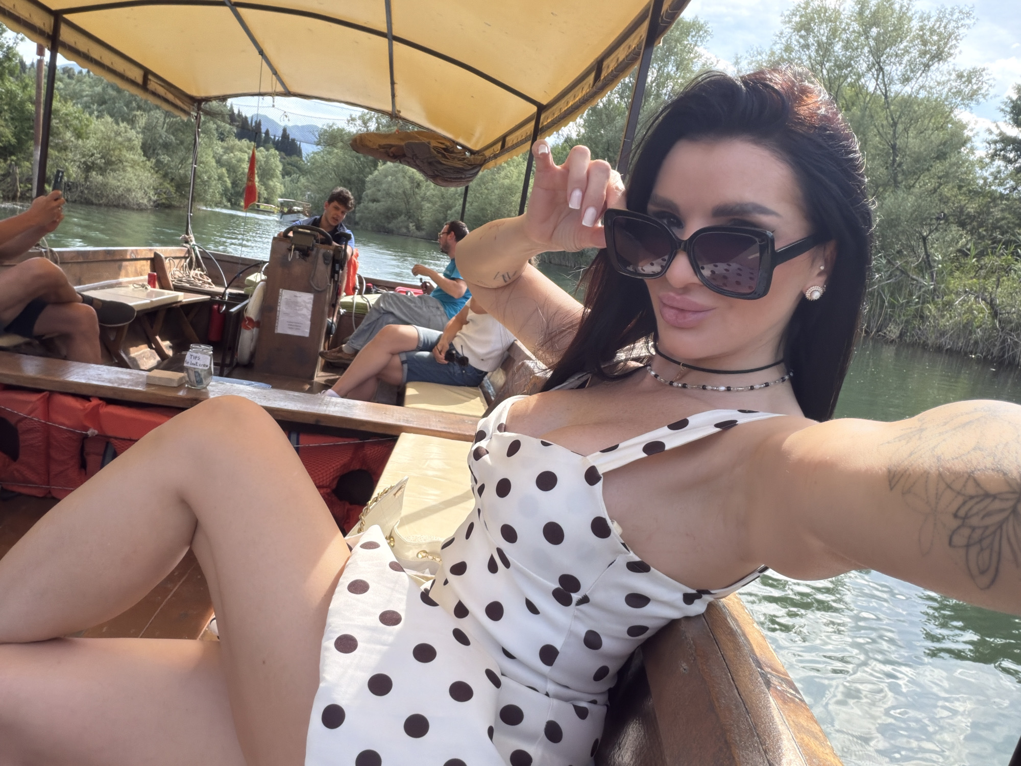 The image captures a selfie taken by a woman with dark hair, wearing large sunglasses and a white dress with brown polka dots, on a boat. She is posing with her hand near her face. In the background, there are other passengers on the boat, including the boat operator and other seated individuals. The boat has a covered top, and the surroundings show a river bordered by lush green trees. This is likely near Boljevići, Montenegro, given the presence of boats, rivers, and greenery typical of that region.