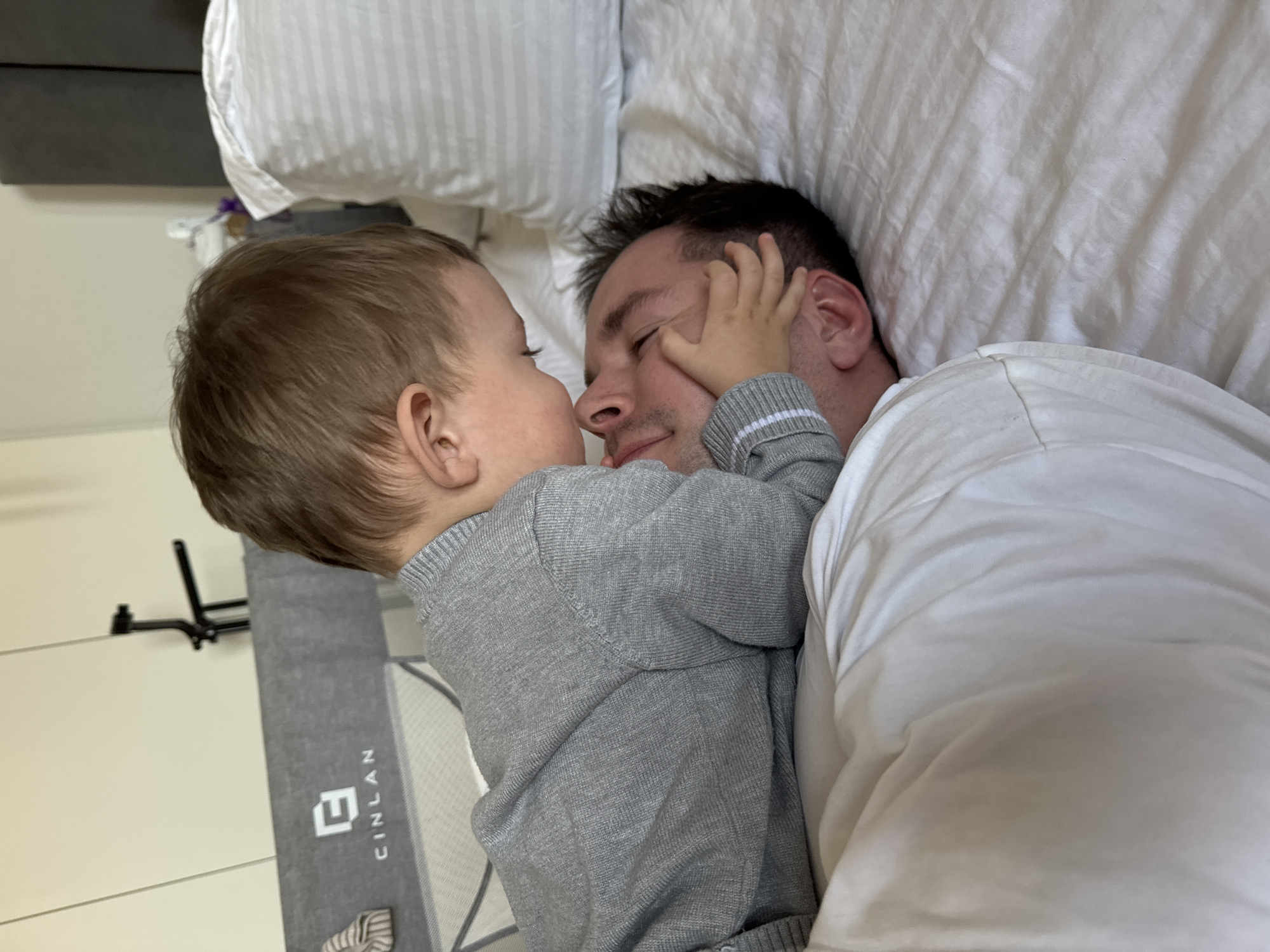 A man and a young child are lying on a bed in what appears to be a residential setting. The man is lying on his back, covered by a white shirt and white bedding. The child, wearing a gray sweater, is positioned beside him, face-to-face, with one hand touching the man's face. The child has light brown hair. A pillow with striped bedding is visible behind the man's head. A wall is visible on the left side of the frame, adjacent to a gray floor mat or play mat that reads "CINLAN" vertically. A black mounting bracket is attached to the wall above the mat. The specific location is not discernible from the image, as the picture only shows an interior. No location is visible from the image to place it in Odintsovo, Russia.