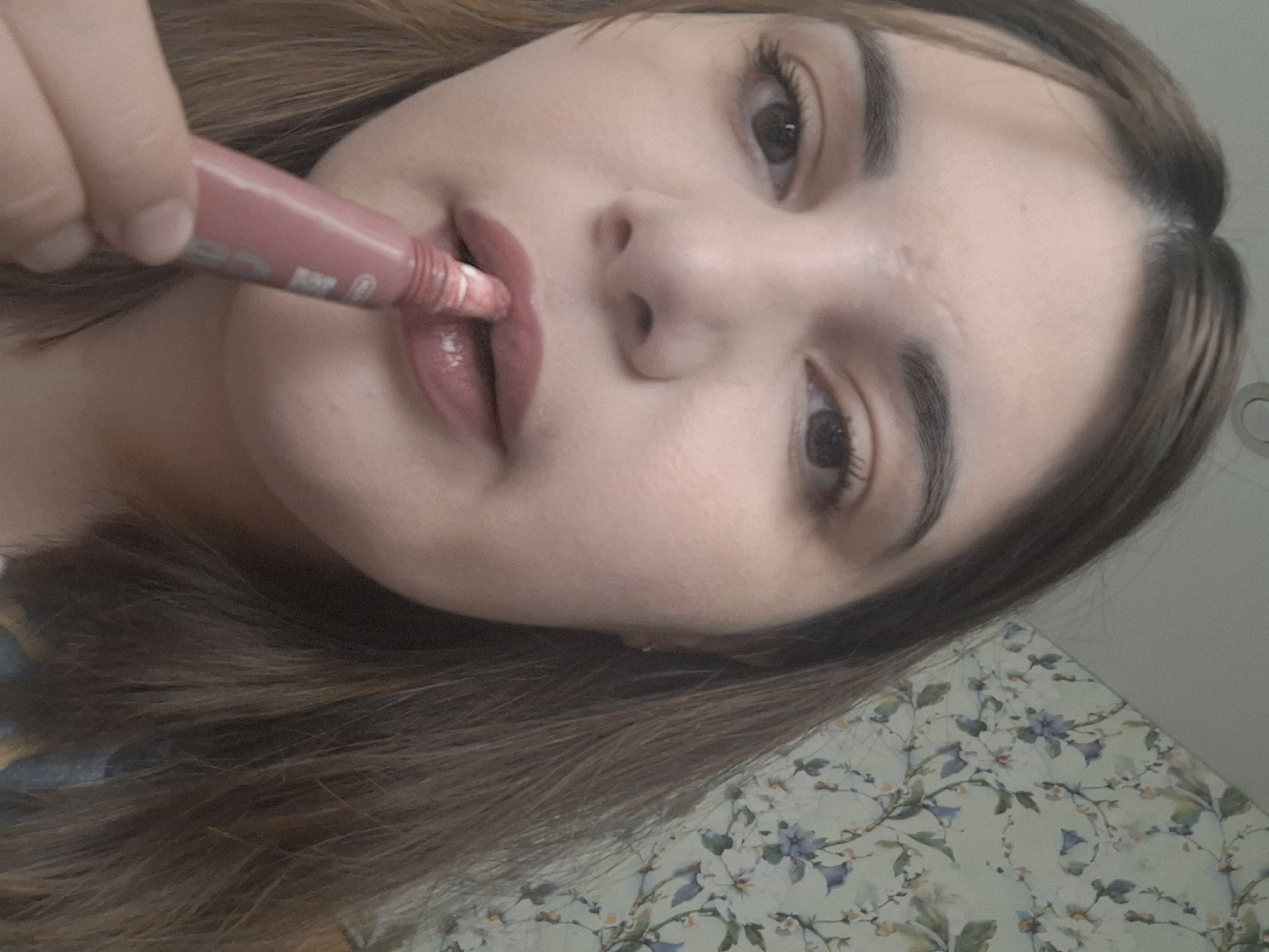 A fair-skinned female is applying a tube of lip gloss to her lips. She has brown eyes, dark eyebrows, and brown hair. The background is a floral pattern. The location of the image is not discernable from the photograph and no other information is available.
