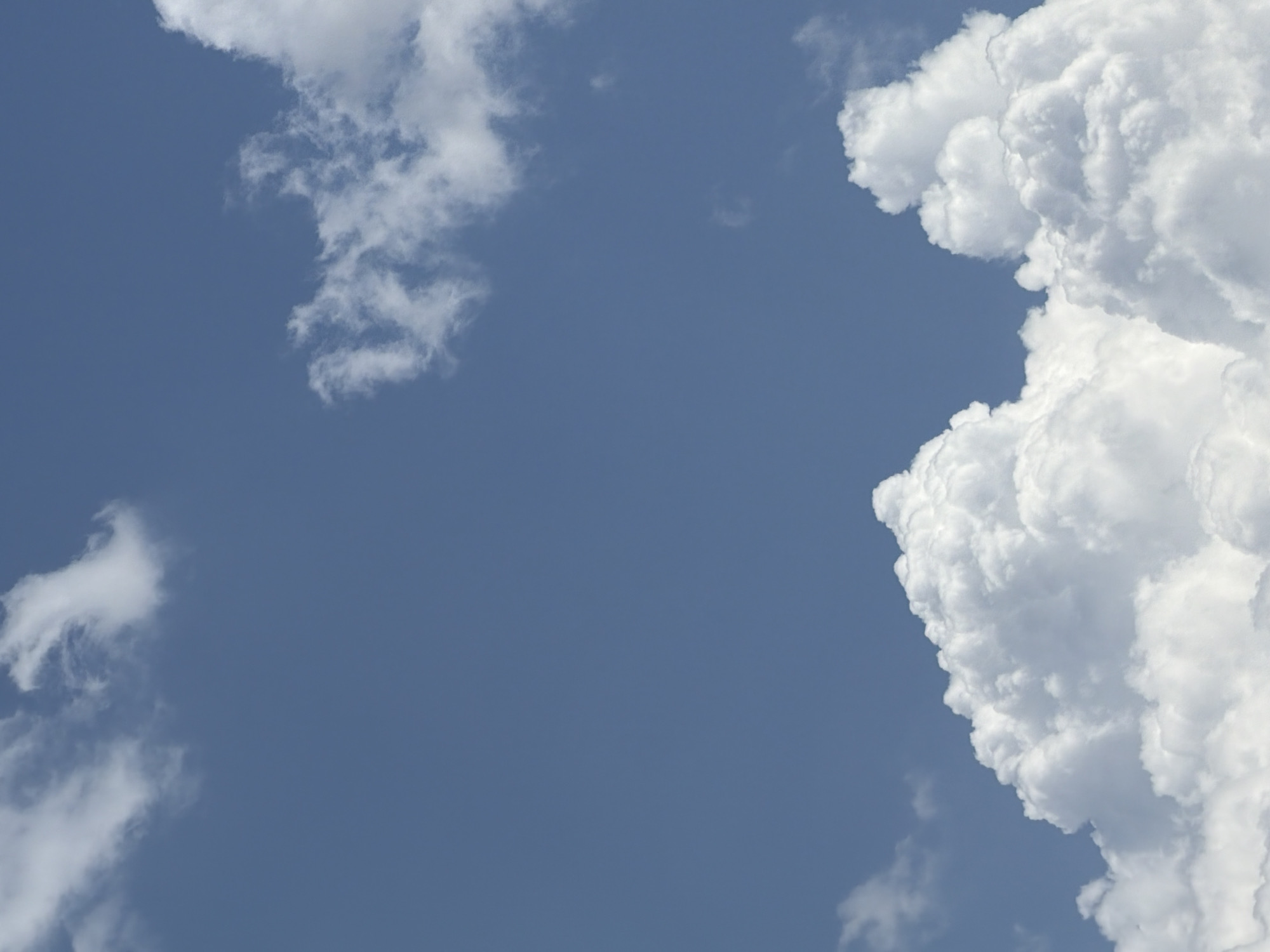The image depicts a bright blue sky with several white clouds of varying sizes and densities. In the lower portion of the image, large cumulus clouds dominate the scene, appearing fluffy and voluminous. Higher up, wispy cirrus clouds are visible, suggesting a layered cloud formation. The lighting indicates it is daytime, likely mid-morning or afternoon, with the sun illuminating the clouds from above. The environment is entirely the sky, with no discernible ground or city elements present. There are no people or objects visible in the frame. The overall impression is of a clear and partly cloudy day.