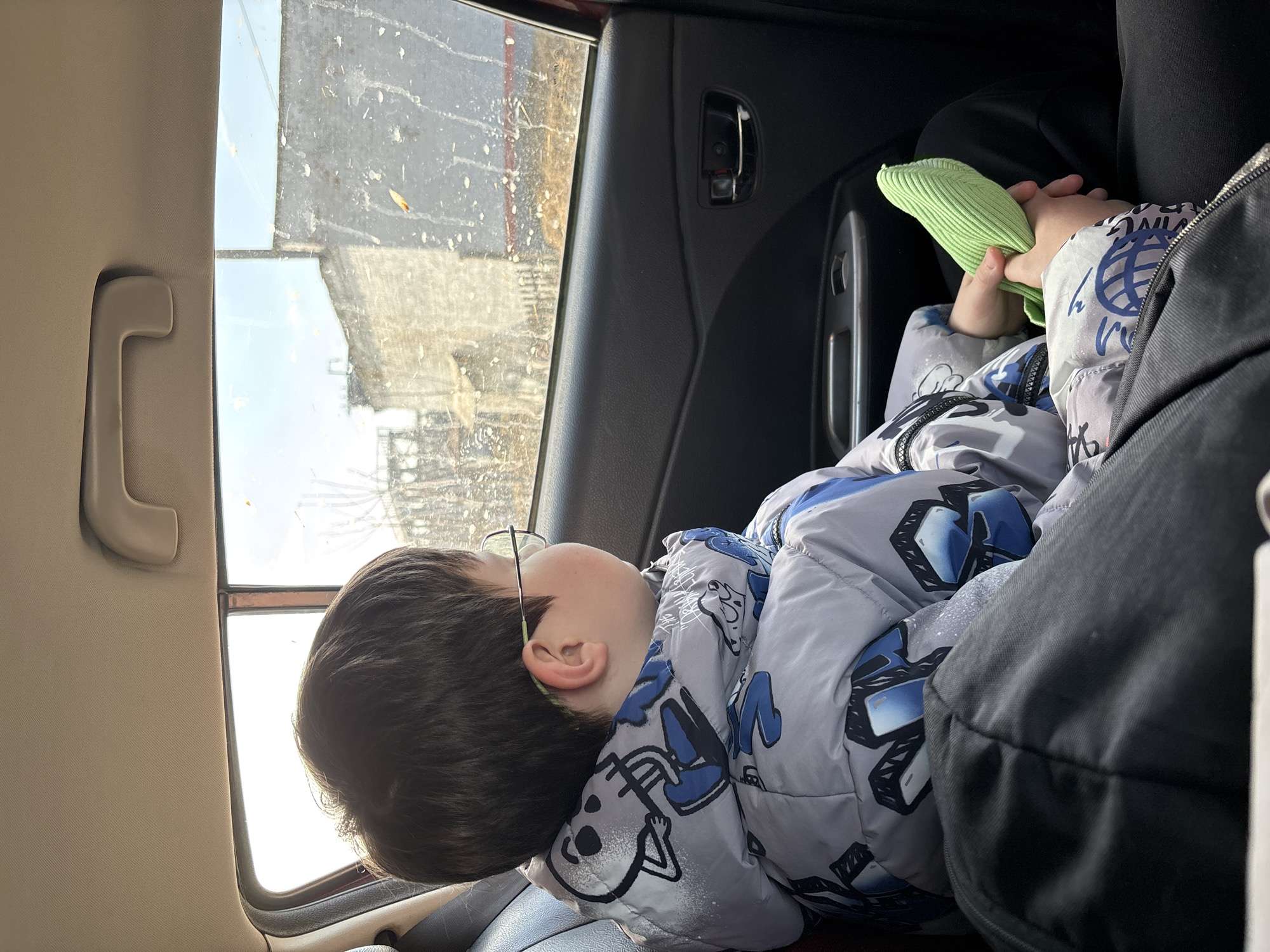 A child with dark hair and eyeglasses rests in the backseat of a car. The child wears a gray and blue patterned jacket and holds a light green ribbed item in their hands. Outside the car window, a cityscape is visible, with buildings and a cloudy sky, potentially indicating the location of Чепца, Russia. The car's interior is visible, including the door panel, handle, and window.