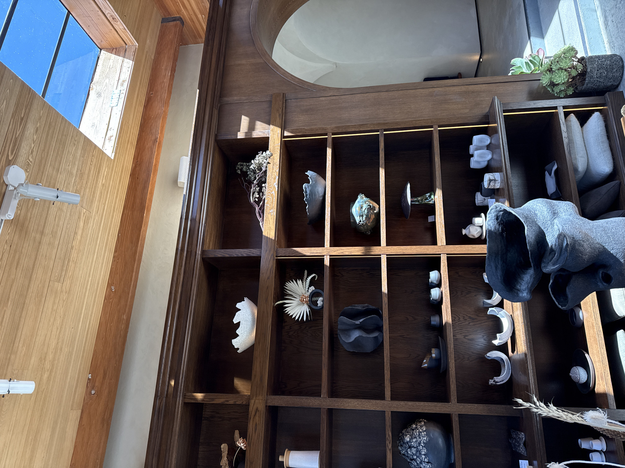 This image shows an interior view of a room in Los Angeles, California, featuring a large wooden shelving unit filled with various decorative objects. The shelves hold an assortment of pottery, sculptures, plants, and other artistic items. A window is visible to the left, with the natural light illuminating the objects. The overall atmosphere suggests a curated, artistic space.