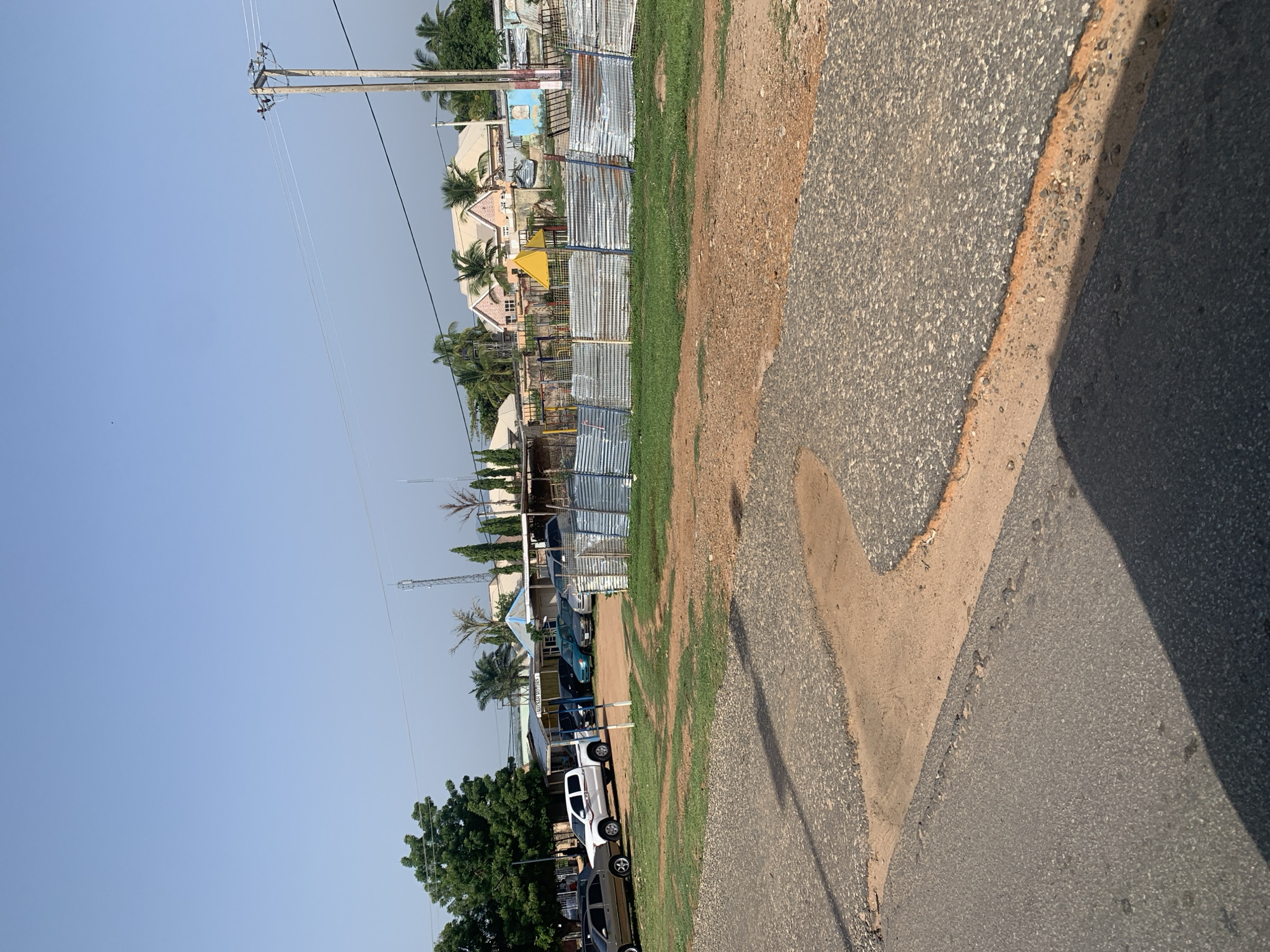 The image depicts an outdoor scene in a somewhat rural or suburban area, with a clear blue sky. In the foreground, there is a paved road with visible patches of dirt and sand. Beyond the road, a grassy area is present, leading to a row of parked vehicles. These vehicles include a white pickup truck, several sedans of various colors (brown, black, blue, green, and silver), and a black SUV. Behind the vehicles, a corrugated metal fence runs horizontally across the scene. Further in the background, a few buildings are visible, some appearing to be single-story structures with light-colored roofs, possibly houses or commercial establishments. There are also trees, including palm trees and other deciduous trees, scattered throughout the landscape. A power pole with wires is situated on the right side of the image. The overall impression is a somewhat arid environment with signs of development and activity.