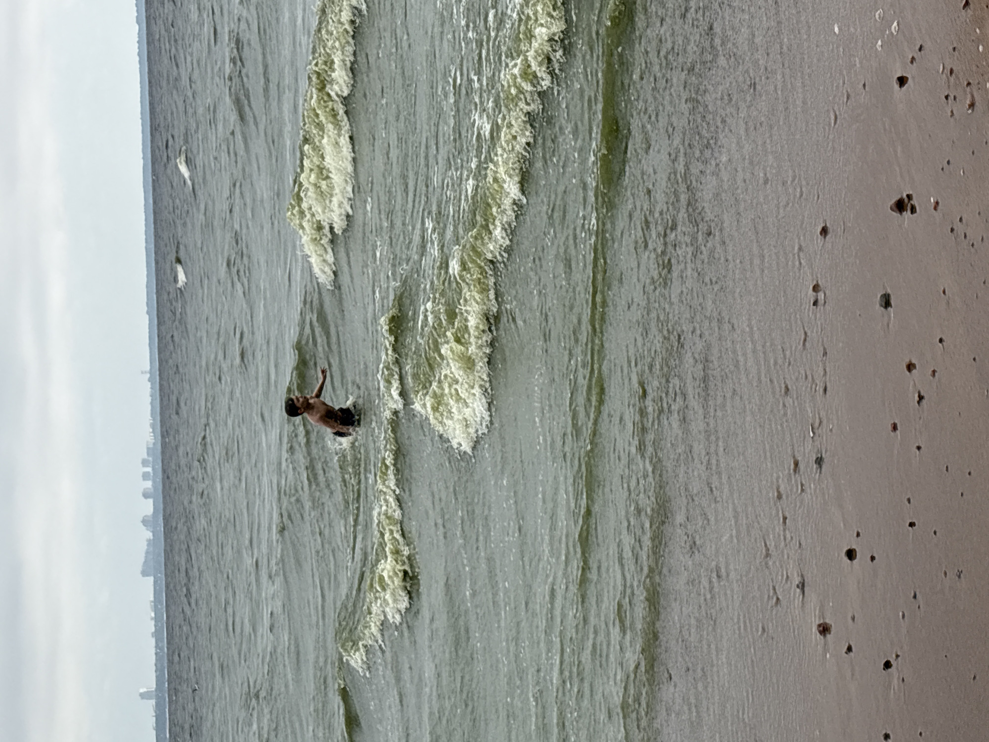 The image depicts a person in the water near a beach. The person appears to be a male, with dark hair, and is partially submerged in the water. They are facing away from the camera and their arms are raised. The water is wavy, with some foam on the crests of the waves. The beach is sandy and appears to have some small pebbles or shells on it. In the background, a hazy cityscape is visible on the horizon, suggesting an urban or semi-urban coastal location. The sky is overcast, indicating a cloudy or possibly dull weather day. The overall scene suggests a casual recreational activity, likely swimming or playing in the ocean. The time of day is not clearly indicated, but the diffused light suggests it could be daytime. There is no visible text in the image. The location context provided is Solnechnoye, Russia.