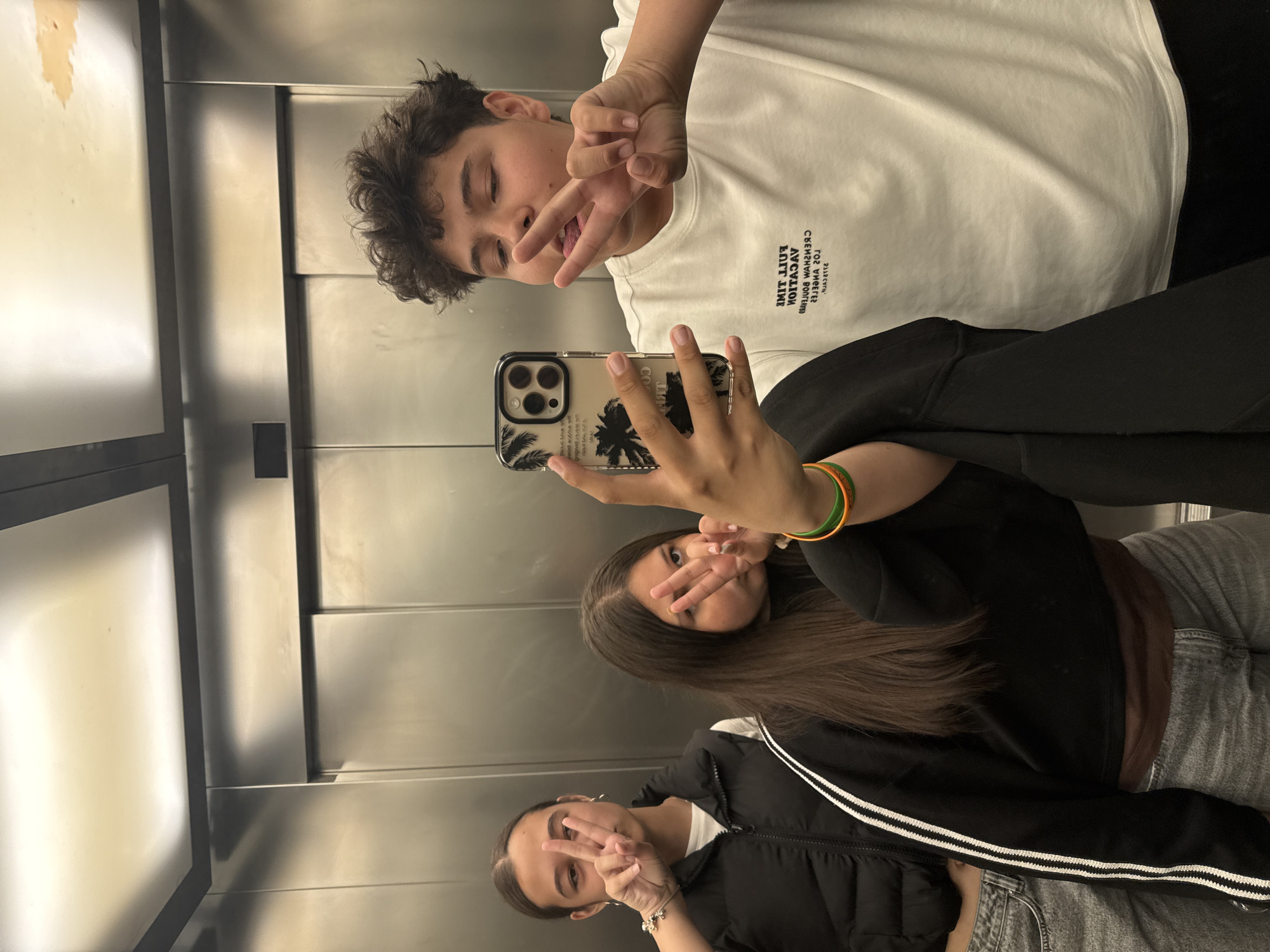 Three individuals are taking a selfie in what appears to be an elevator. The elevator's interior is characterized by stainless steel panels. The person at the top is a young male with curly, dark hair, wearing a white shirt. The individual in the middle has long, straight brown hair, wearing a black jacket. The third individual is a young female wearing a black puffer jacket with white stripes. All three are making peace signs with their hands. The person in the middle holds a smartphone with a clear case featuring palm tree designs. The location is Unknown, Unknown.