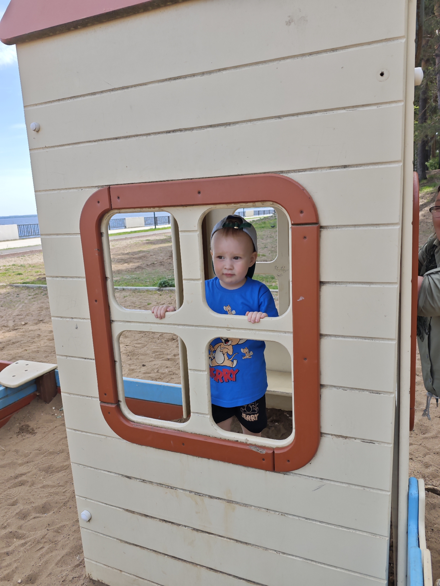 A young child wearing a blue T-shirt and a backwards baseball cap is looking through a window in a playhouse located in a sandy area. The playhouse has a red-brown trimmed window. In the background is a body of water and a cement barrier with a railing. The scene is in Chaikovsky, Russia.