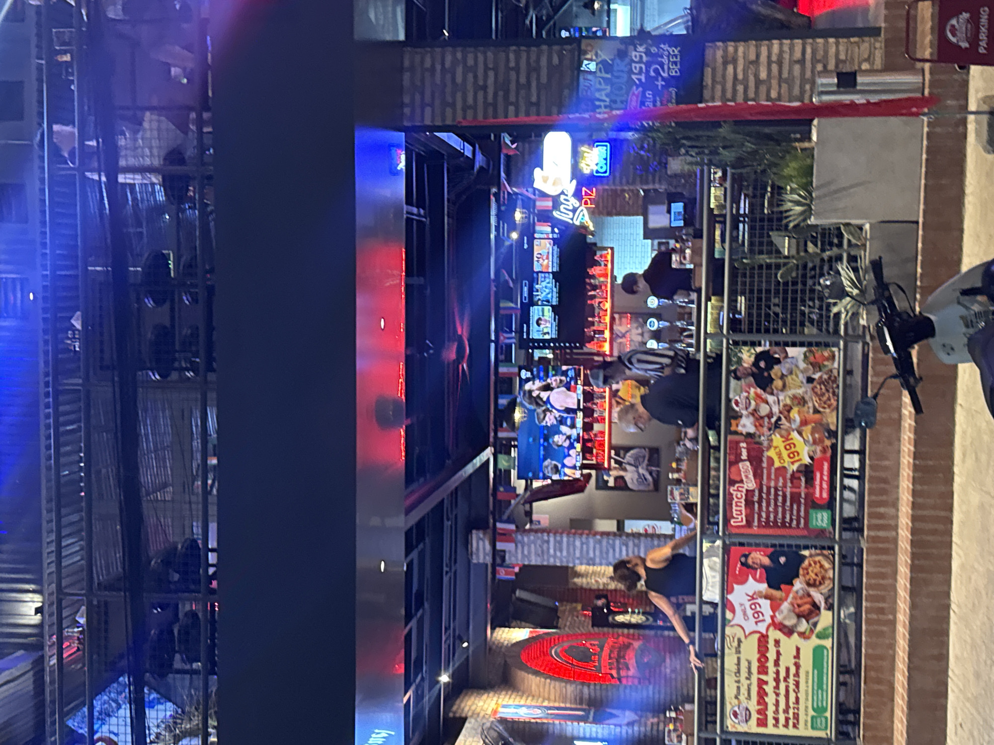 The image captures a bustling outdoor dining or bar area, likely in Bac Nha Trang Ward, Vietnam, given the presence of Vietnamese currency denominations and typical Southeast Asian street scene elements. In the foreground, a partially visible motorcycle is parked on a sidewalk. Above the sidewalk, there is a railing adorned with two large advertising posters. The poster on the left promotes "HAPPY HOUR" with "Pizza & Chicken Wings" for "ONLY 199K". The poster on the right advertises a "Lunch COMBO for 2" also for "ONLY 199K".
Behind the railing, a bar area is visible, with bottles of alcohol neatly arranged on shelves. Several people are present in this area, including a woman in a black tank top facing away from the camera, appearing to lean on the railing. To her right, a man in a black shirt is working behind the bar. Further to the right, two men are seated at a table, one appearing to be an older gentleman with glasses. The background reveals a multi-level structure with exposed brick and metal framing, illuminated by red and blue neon lights and television screens displaying sports or entertainment. A prominent neon sign reading "Ing PIZ" is visible. The overall atmosphere suggests a lively evening entertainment venue.