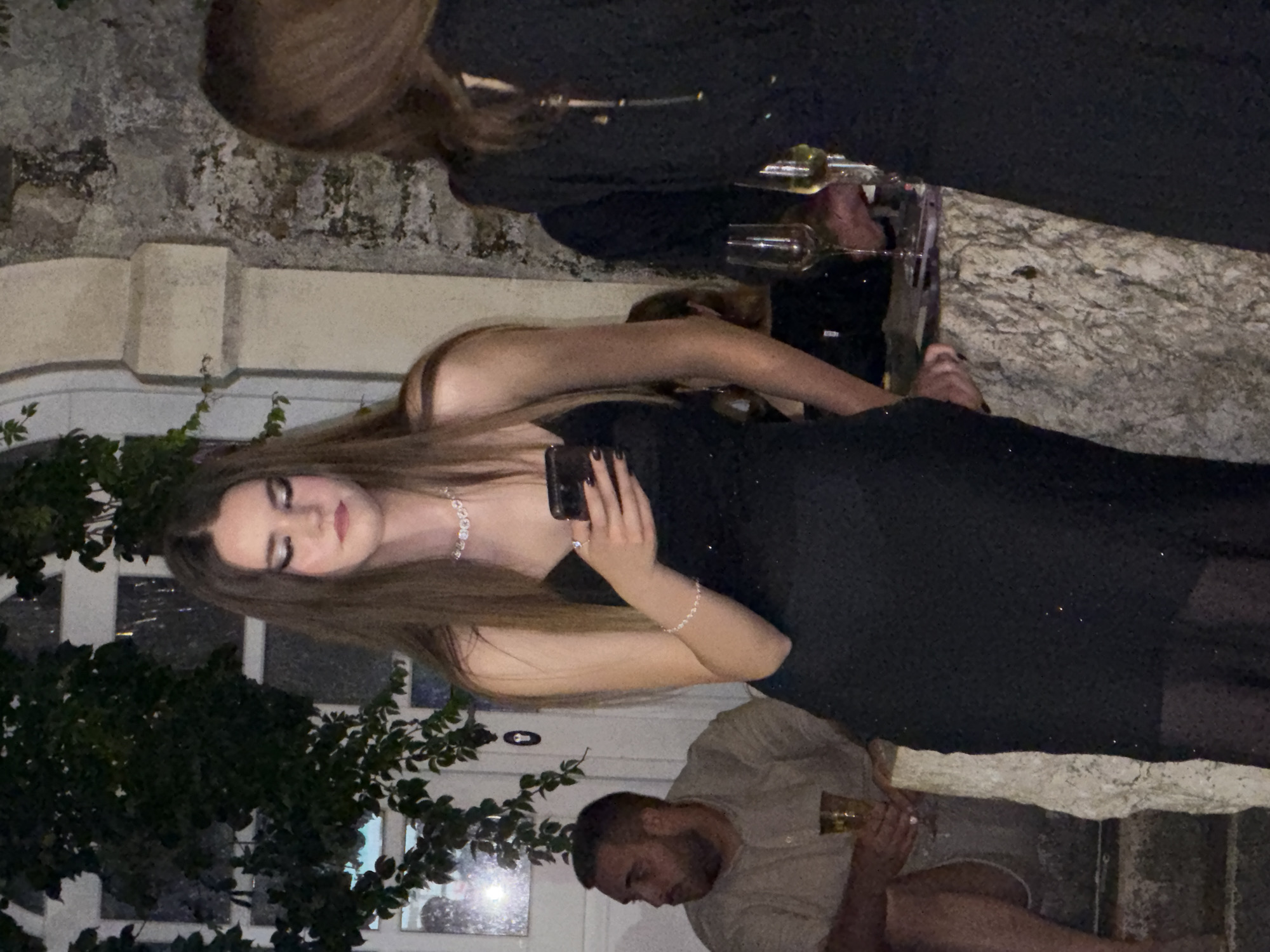 The image shows a group of people at an outdoor event in Budva, Montenegro. The primary subject is a young woman in a black dress and diamond necklace, holding a phone. She appears to be looking at the phone screen with a neutral expression. To her right, another woman in a black dress is visible, holding a glass. In the background, a man in a light-colored shirt is seen holding a drink, with his head bowed. The scene is illuminated by artificial lighting, suggesting it is nighttime or evening. The setting appears to be a courtyard or patio, with stone walls and lush greenery. The overall mood seems relaxed and social, with people enjoying drinks and each other's company. No visible text is present in the image.