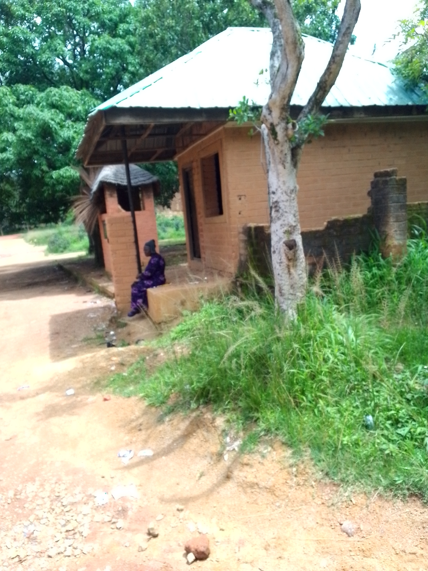 The image depicts an outdoor scene with a small brick building and surrounding vegetation. A person wearing a purple, patterned garment is seated on a low, concrete structure to the left of the building. The building has a light-colored, corrugated metal roof supported by wooden beams. A narrow opening, possibly a doorway, is visible on the front of the building. To the right of the building, a weathered brick column stands. In the foreground, the ground is a mix of reddish-brown soil and small stones, with patches of green grass and weeds. A thin, textured tree trunk with sparse leaves is positioned in front of the building. The background consists of lush green trees, suggesting a natural or semi-rural environment. The scene appears to be located in the city of Jos, Nigeria, characterized by its typical architecture and vegetation.