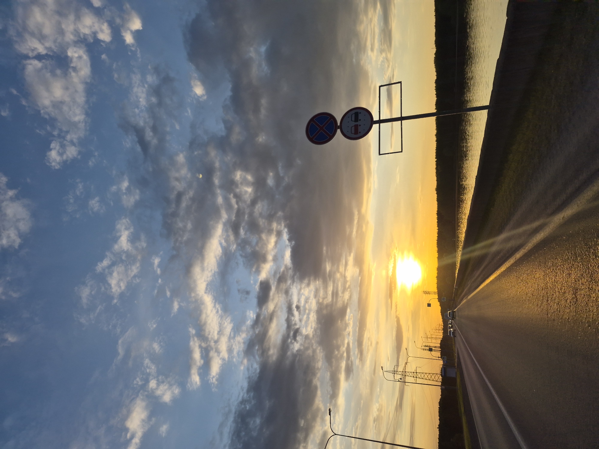 The image shows an eastward-facing view along a highway during sunset in Noviy, Russia. The road extends towards the horizon, reflecting sunlight. Several cars are on the road. Traffic signs are visible on a pole, indicating restrictions on stopping and vehicles carrying dangerous goods. Electrical towers are lined along the roadside. The sky is partially cloudy, with blue tones on the left and sunset colors dominating towards the right. The road is bordered by foliage and possibly a body of water on the right side.