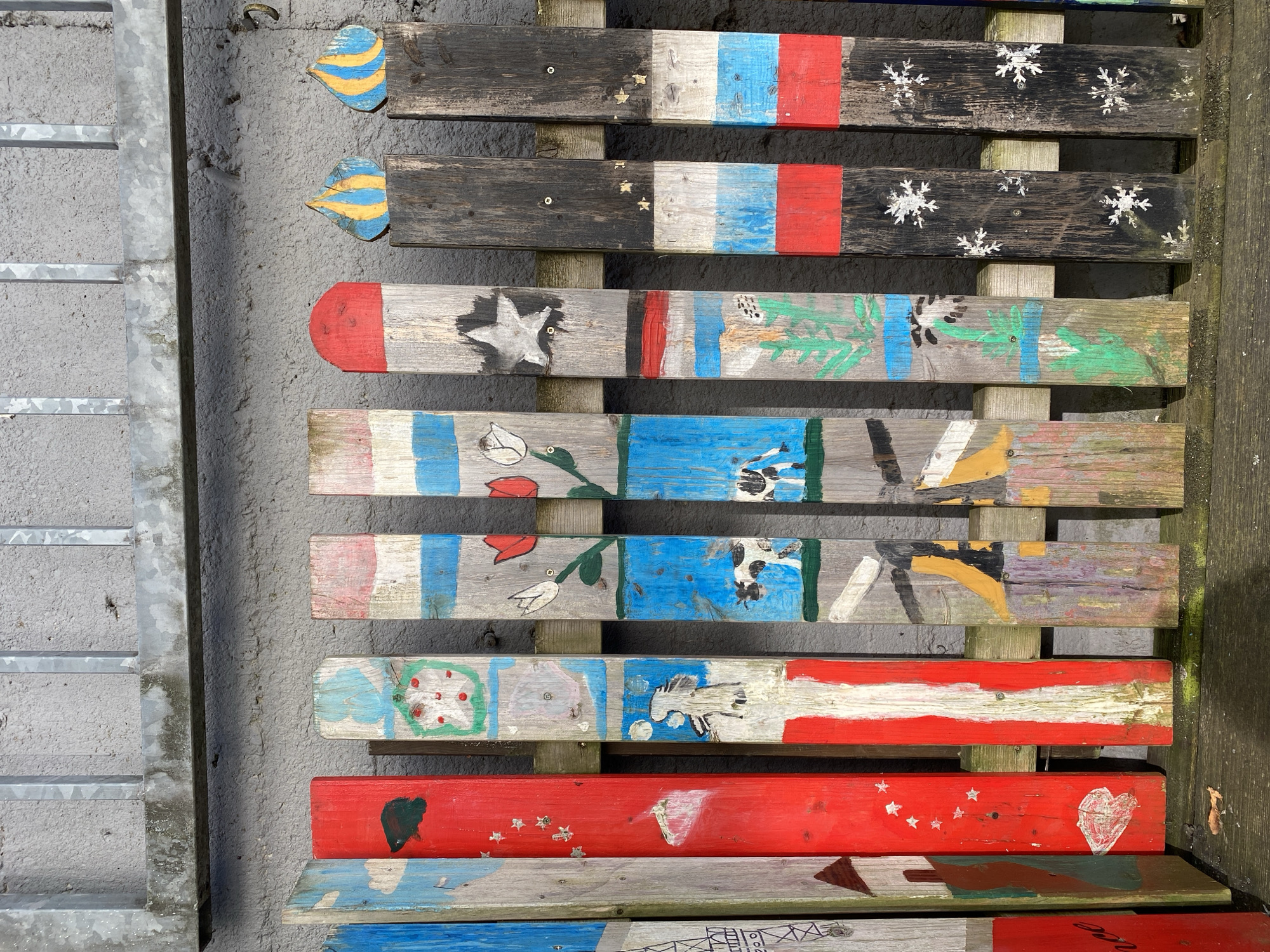 The image shows a close-up, eye-level view of a rustic wooden fence with various colorful paintings on its slats, situated outdoors against a rough concrete wall. The fence is comprised of vertical wooden planks, some of which are decorated with simple, folk-art style paintings.
The painted designs vary widely across the slats. From left to right, one plank appears to depict a building with a red roof, possibly a house. The next plank is solid red, adorned with white stars and a white heart near the bottom. Following this is a plank painted with vertical red and white stripes, with some apparent wear. Another plank features a depiction of a red apple in a green circle and further down, a black and white cow. Next to it, a plank shows two tulips, one red and one white, on green stems, with a blue background. Further to the right, a dark wooden plank is painted with a white star. The subsequent planks are dark brown or black, with patterns of white snowflakes. At the very right, two slats are topped with decorative elements resembling flame or teardrop shapes, painted in yellow and blue stripes. Horizontal wooden support beams are visible behind the painted slats. The concrete wall behind the fence is rough in texture and light gray in color, with some visible dirt or staining. Above the concrete wall, a section of a metal fence or railing with vertical bars is visible, also showing signs of wear and discoloration. The ground in front of the fence appears to be wooden planks, possibly part of a bench or walkway, with some fallen leaves. The lighting suggests it is daytime, with sunlight casting shadows on some of the fence slats. There are no people or animals visible in the frame. The overall impression is one of a decorative, perhaps children's art project, integrated into an outdoor setting.
This fence is located at the Albert-Schweitzer-Schule in Altenkirchen, Germany.
