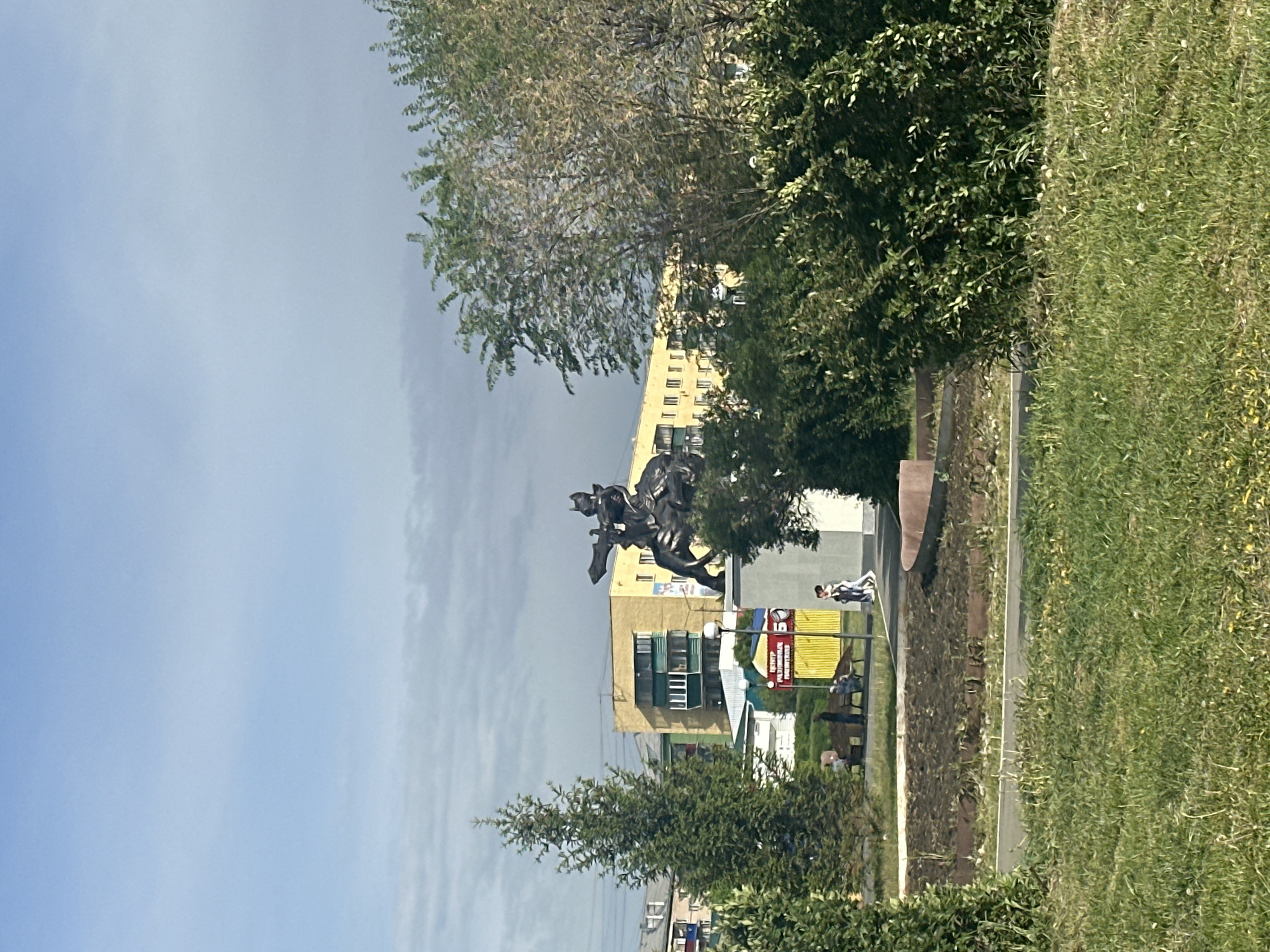 A landscape-oriented photo shows a bronze statue of a mounted warrior on a raised platform in front of a tall, light-yellow building. The statue is situated on the side of a road, with a narrow sidewalk and grass on the right side of the road. Two people walk away from the viewer along the sidewalk. To the right of the statue is a red, white, and yellow sign behind a stone wall. Trees are scattered throughout, partially obscuring the building and statue. The sky is a pale blue with some clouds.
