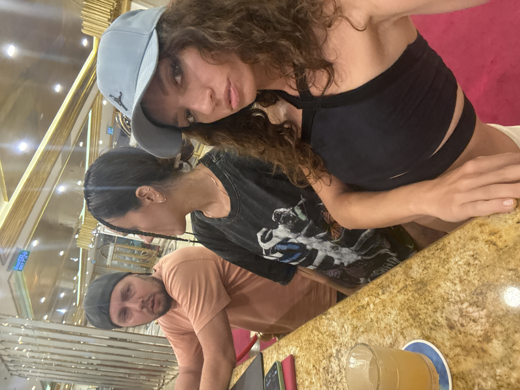 A poorly angled close-up photo showing three people seated at a bar. The primary subject, a woman with curly hair, wears a light blue baseball cap and a black crop top. Beside her, another woman with dark hair in braids wears a graphic t-shirt. A man in a tan shirt and grey cap is on the left, partially obscured. A glass of liquid is visible on the bar. The background features a glitzy, gold-toned interior. An exit sign in Vietnamese is visible. No specific mention is given that this is in Đà Nẵng, Vietnam.