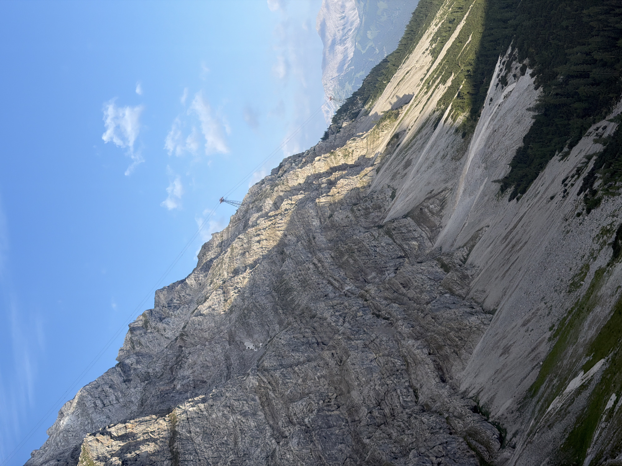 The image depicts a steep, rocky mountain slope under a clear blue sky with scattered clouds. A cable car system with two towers and wires traverses the mountain. The lower part of the slope is covered with scree and sparse vegetation, transitioning into a dense evergreen forest at the base. In the background, more distant mountains are visible, some with patches of snow. The lighting suggests it is either early morning or late afternoon, with a distinct shadow line cutting across the slope. The scene is a natural mountain landscape, likely a tourist destination given the presence of the cable car. This location is consistent with the Zugspitze area near Grainau, Germany, which features similar mountainous terrain and cable car infrastructure. There are no people or specific actions visible in the image.