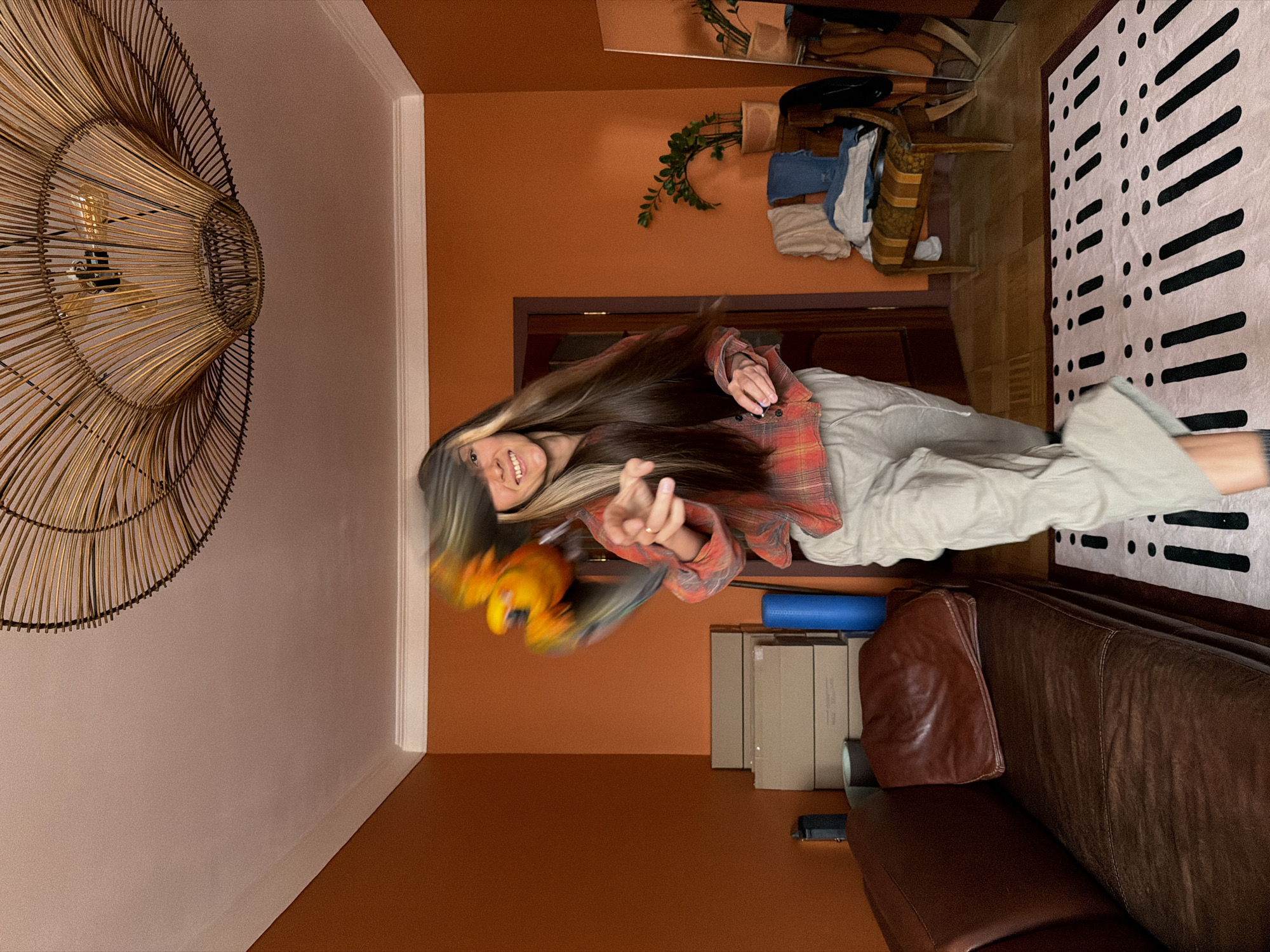 A room with orange walls and a ceiling that transitions from peach to white, dominated by a large, intricately woven rattan light fixture affixed to the upper left. A young woman is in the center; she has long hair and wears a red plaid shirt and grey pants. She is smiling and making a hand gesture. A yellow and green parrot is blurred and in motion near her left hand. Behind the woman, a doorway is visible. In the background there are some shelves or a table with items and a potted plant. To the right, a black and white patterned rug is on the floor, alongside a brown leather sofa. Stacks of cardboard boxes are visible on the floor near the sofa. There is no evidence to indicate a location in the city of Khimki, Russia.