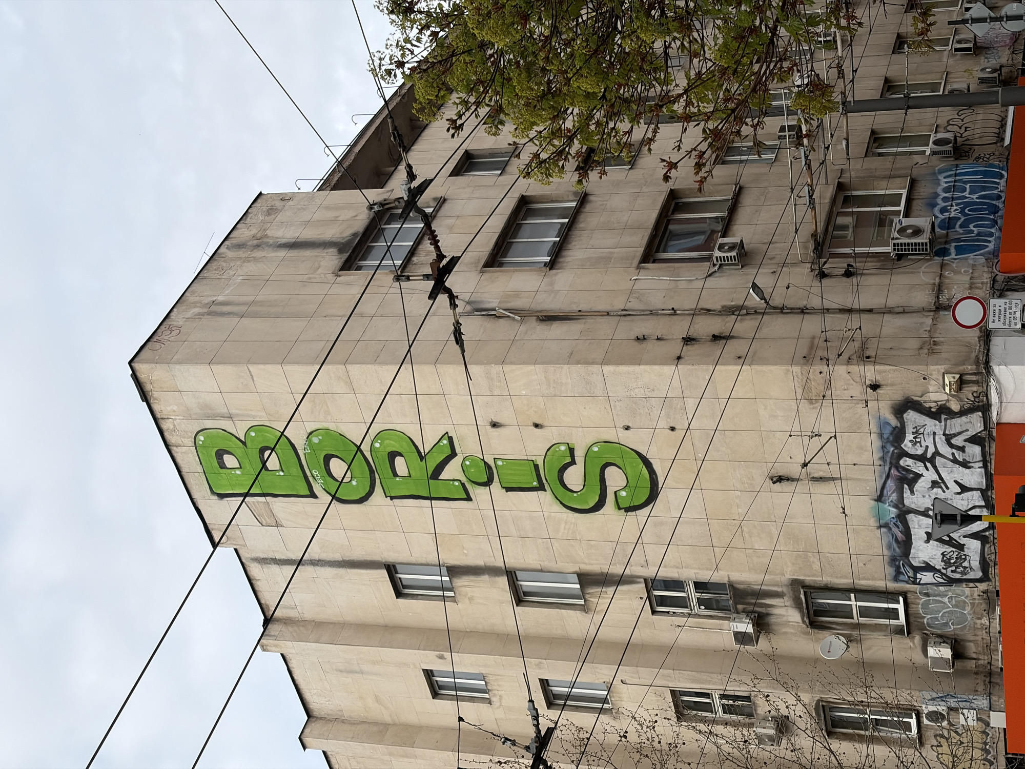 An urban scene in Sofia, Bulgaria, features a multi-story building facade with a worn, light-colored tiled or stone exterior, seen from a low-angle perspective. The camera is rotated clockwise, making the building appear on its side. When viewed upright, the building rises against an overcast, light gray sky.
The most prominent feature is a large, vibrant green graffiti tag spelling "BORIS" (or similar letters, considering rotation and OCR) across the upper-middle section of the building. Smaller "ECO" text is visible vertically near the 'R'. To the right, a large, abstract graffiti piece in blue, white, and gray tones covers a significant portion of the facade. Numerous black electrical or communication wires crisscross the entire front of the building, connected to various anchors and poles, highlighting the dense urban infrastructure.
The building features multiple rectangular windows across several floors. Many windows have external air conditioning units mounted beneath or beside them. A small white satellite dish is also visible. Tree branches with light green foliage, indicative of spring, frame the top and upper-right corner of the image.
On the far right, a circular red and white regulatory sign (likely "No Stopping" or "No Entry" variant) is present. Below it, a white rectangular sign displays Cyrillic text, which the OCR identifies as: "Не важи за живущи и обслужване на обекти от 00.00 40 057-са". This translates to "Not valid for residents and servicing of objects from 00:00 to 05:00", indicating specific time restrictions. The overall impression is a bustling urban environment, typical of a city setting, with elements of street art and visible utility infrastructure.