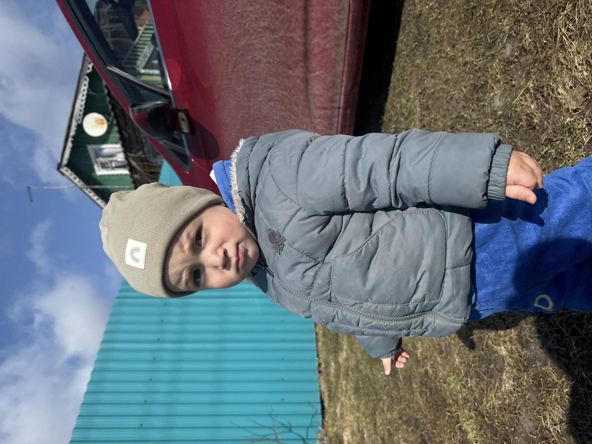 A child wearing a khaki beanie, gray puffy jacket, and blue pants stands outdoors. A red car is in the background, partially obscuring a green house. A blue corrugated metal structure is visible to the left, and the ground is covered with grass and dirt. The sky is visible in the upper left corner, with light clouds. The location is Unknown, Unknown.