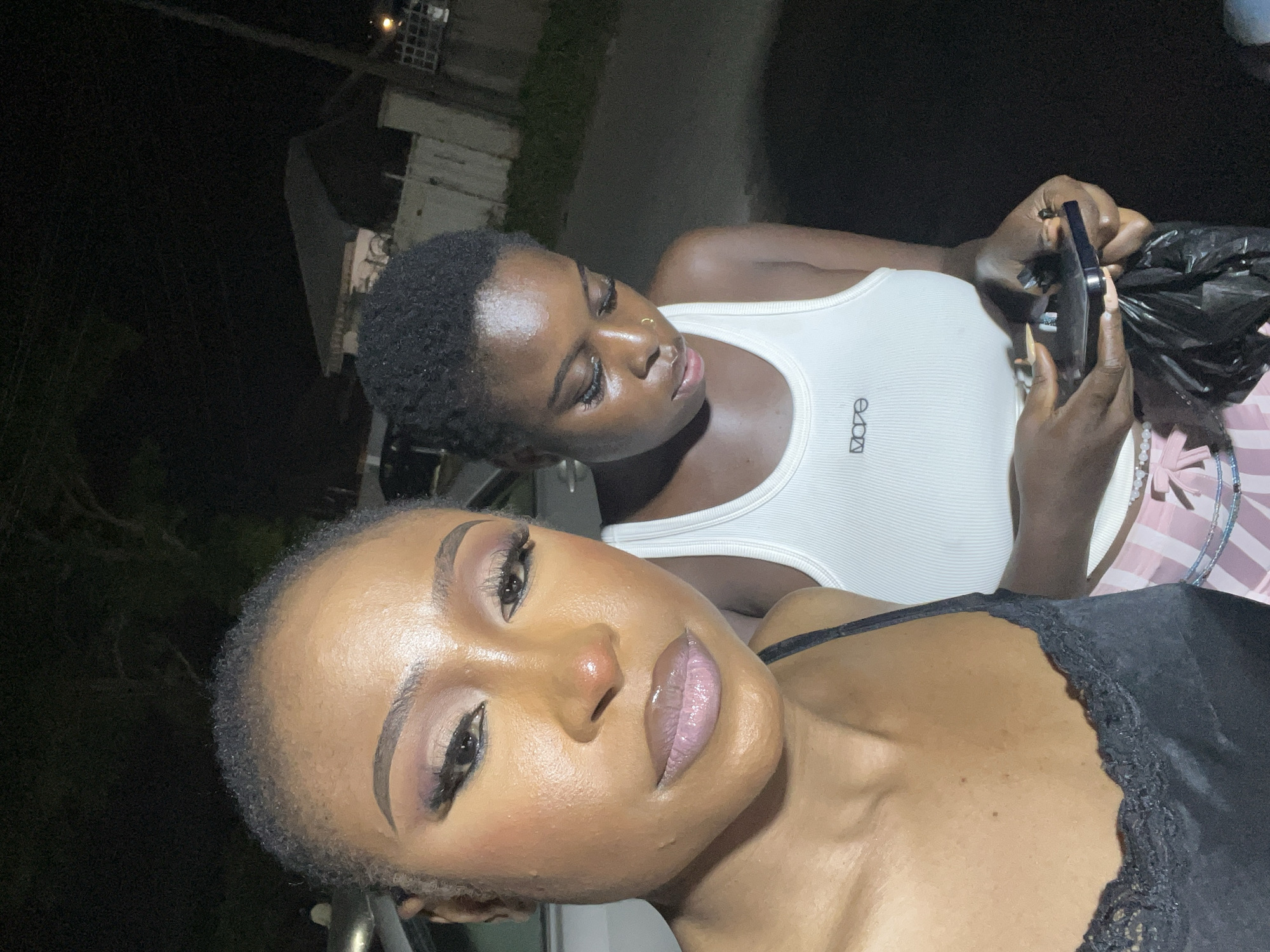 Two women are standing outdoors at night in what appears to be an urban environment in Abuja, Nigeria. The woman in the foreground is positioned slightly to the left, facing the camera and looking directly into it. She has her makeup done, including eyeshadow, eyeliner, and lipstick. She is wearing a black tank top with lace trim. To her right and slightly behind her is another woman looking down, focused on something in her hands. This woman is wearing a white ribbed tank top with a small logo on it and what appears to be striped pants. She is holding a black plastic bag and a small dark object in her hands. The background is dark, suggesting it is nighttime, with some indistinct structures and foliage visible. A portion of a vehicle is visible on the far left.