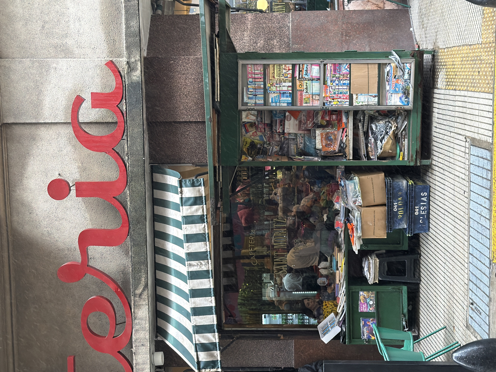 The image shows a street-level view of a corner establishment in Buenos Aires, Argentina. The establishment is "Saint Moritz Cafeteria 1959." A newsstand is positioned outside the establishment's window, stocked with magazines, newspapers, and other goods. A red sign is visible next to the store. The sidewalk is paved with tiles, and stairs lead to another level next to the shop. A green and white striped awning shades the shop window. There are a few people visible in the shop, their faces partially obscured by merchandise and reflections. Crates are stacked atop the newsstand. A small green chair sits by the edge of the newsstand, near the tiled sidewalk.