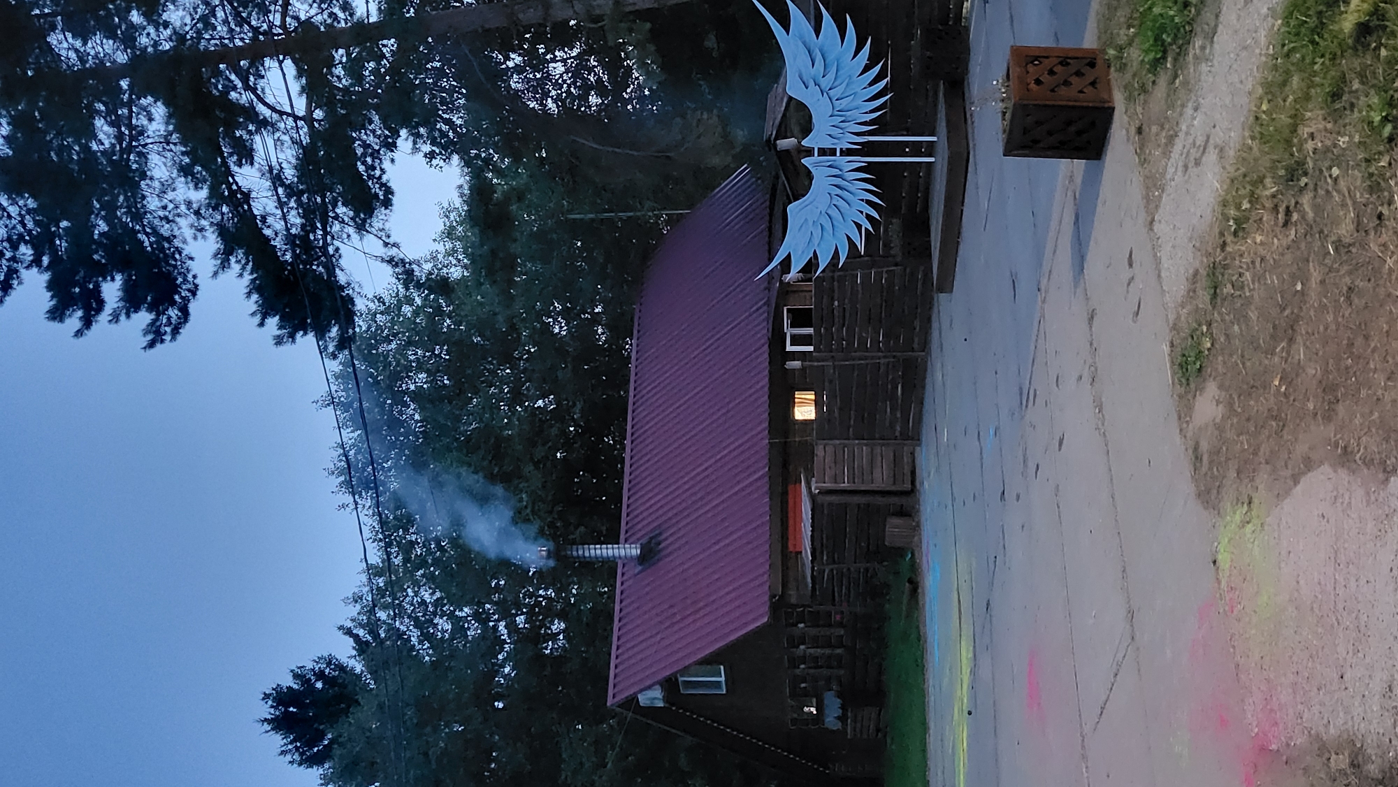 This is a photograph taken in the city of Prikamskii, Russia, at dusk. The scene depicts a rustic wooden house with a dark red metal roof. A metal chimney on the roof is emitting a plume of white smoke. The house is surrounded by a dark wooden fence, and there are several windows visible on its facade. In front of the house, to the right of the image, is a large, white, stylized set of angel wings mounted on a stand. The ground in front of the house is paved with concrete, and there are colorful chalk marks scattered across it, including pink, yellow, and blue. In the foreground, to the right, is a square metal planter with a decorative lattice design, containing some sparse greenery. The background is dominated by a dense forest of trees. The sky is a deep blue, indicating the end of twilight. There are no people visible in the image.