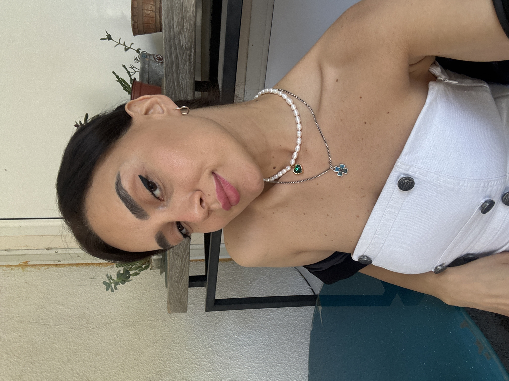 In an outdoor setting, a woman with dark hair is captured in a close-up, high-angle shot. She is wearing a white strapless top and two necklaces, one with pearls and a green heart pendant, and another with a cross pendant. Her makeup includes winged eyeliner and pink lipstick. The background features a textured wall, a shelf with potted plants, and a dark blue surface. The scene appears to be in Tbilisi, Georgia, as indicated by the stucco architecture and the type of greenery.