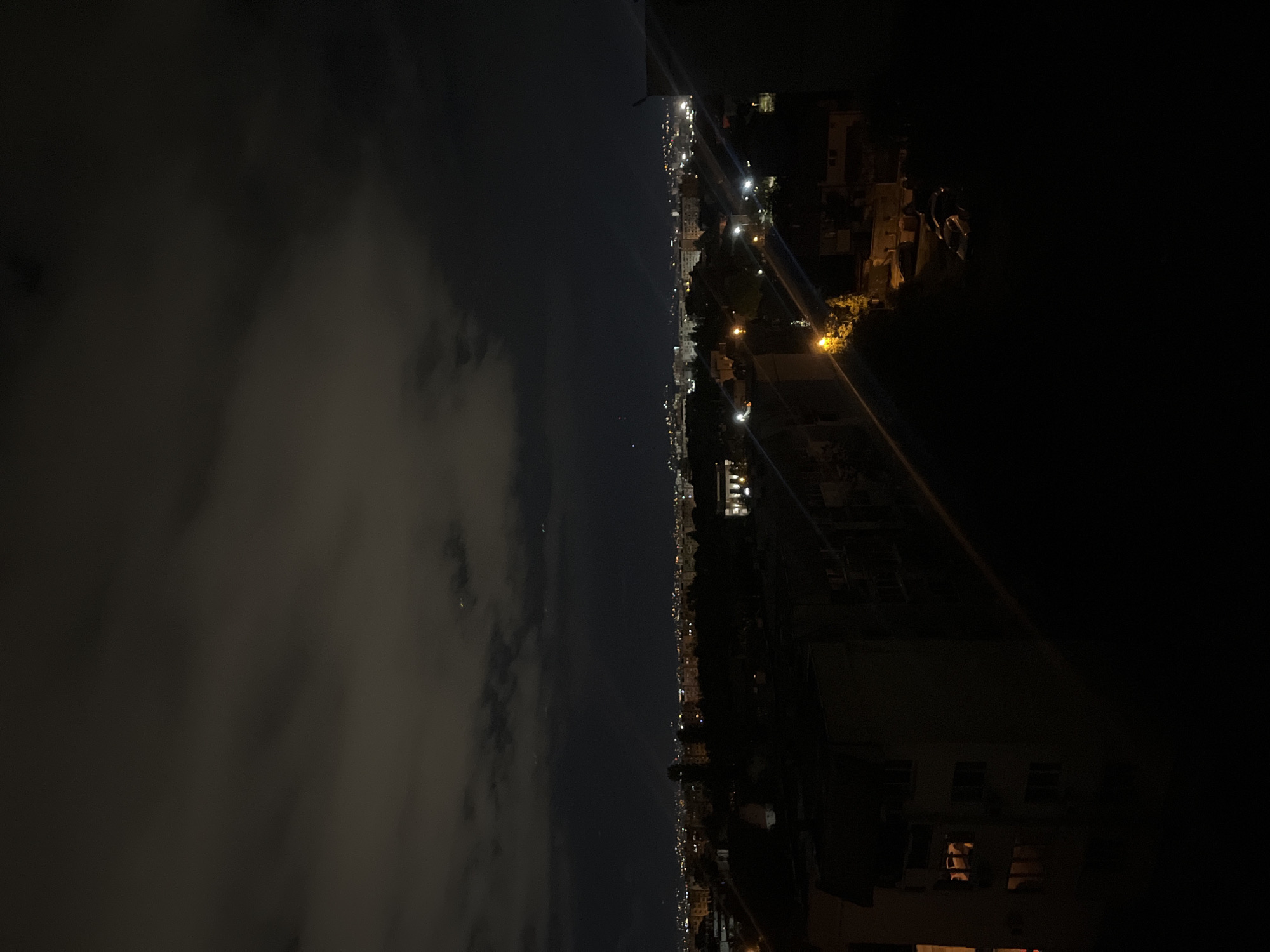 The image depicts a nighttime scene, likely taken from a high vantage point in Sofia, Bulgaria. The left side of the image shows a dark sky with scattered clouds. The center reveals a city skyline illuminated by streetlights and building lights. The right side of the image shows buildings and trees in darkness, with a visible streetlight. It is hard to get specific geographical information without more landmarks.