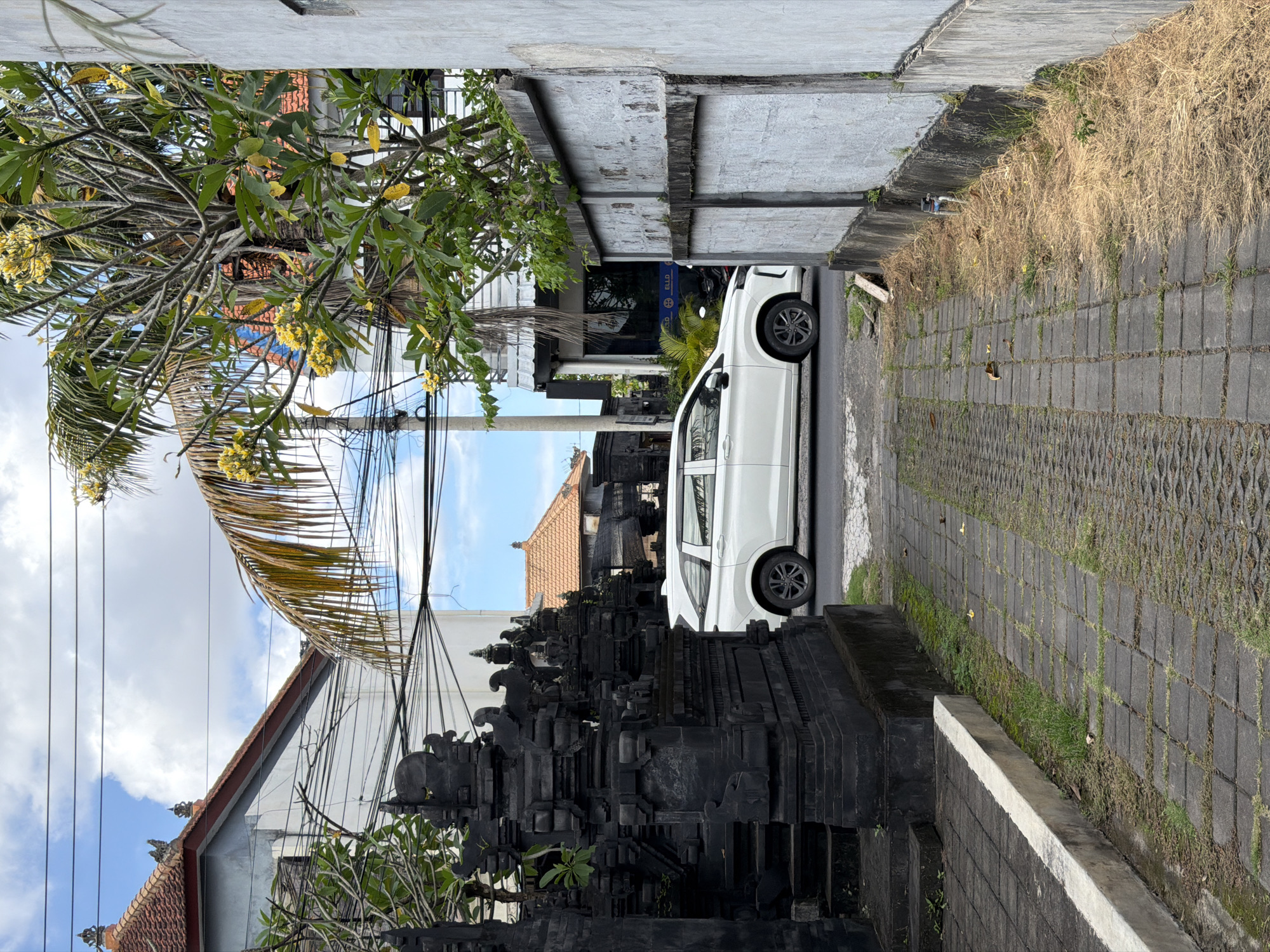 The image depicts a street scene in North Kuta, Indonesia. A white minivan is parked on a narrow road between a traditional Balinese black stone structure, likely a temple or family shrine, and a concrete wall. Paved stone blocks form the road and part of the sidewalk. A tree with yellow flowers partially obscures the view on the left. Overhead power lines crisscross the sky. In the background, buildings and a shop with a blue sign are visible. Dry grass and greenery grow along the edges of the road and sidewalk.