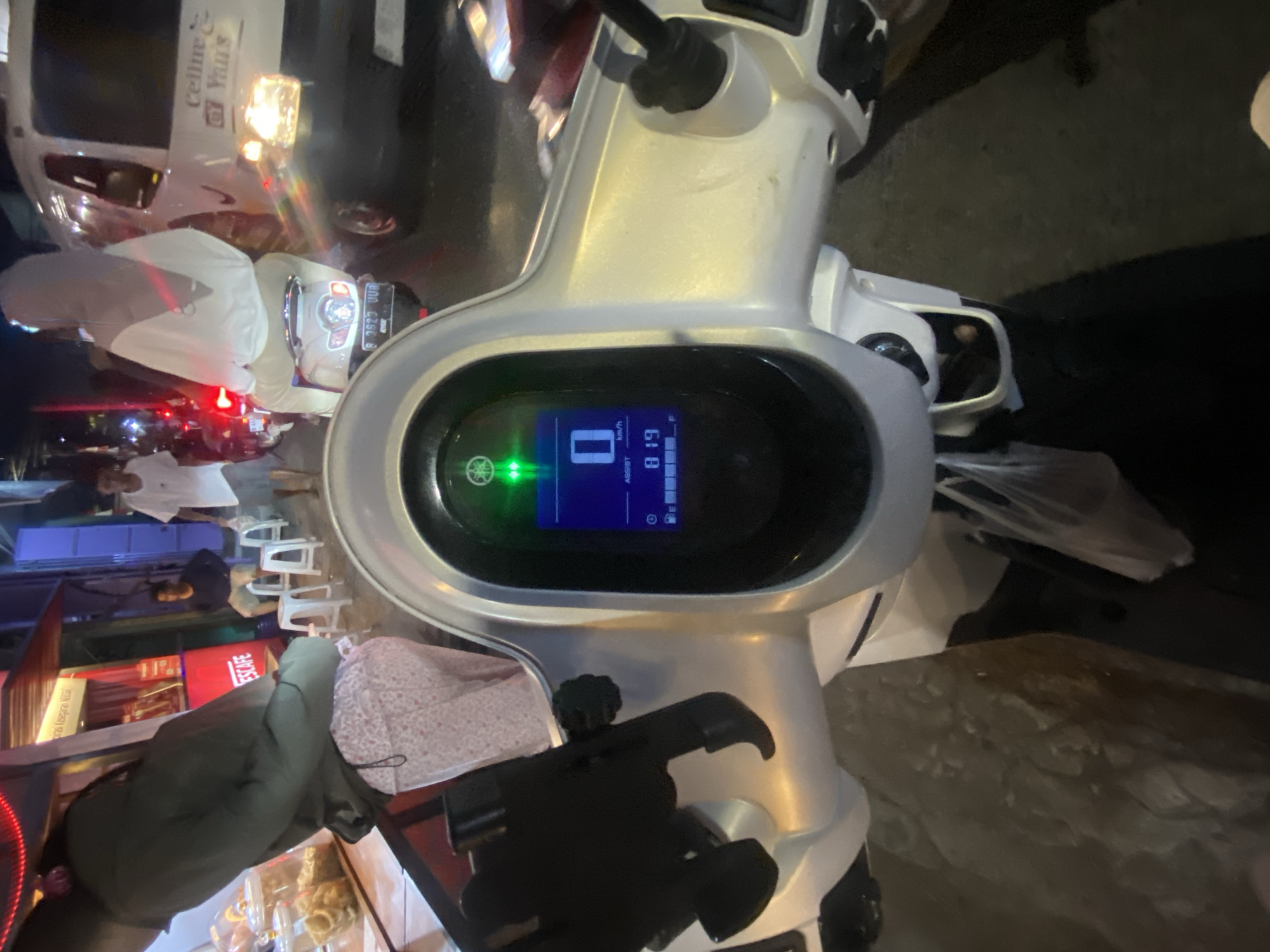 The image shows a first-person perspective from a scooter in North Jakarta, Indonesia. The scooter's digital dashboard displays a speed of "0 km/h" and an odometer reading of "819". Two green indicator lights are illuminated above the display. A phone mount is attached to the handlebars. In the background, a woman wearing a hijab and a white top is riding a white scooter. To the right, a white van with the text "Celline & GY Yura's" is visible, with its headlights on. On the left, a person wearing a green hoodie and patterned pants is standing near a food stall with red and yellow signage. Other people and white plastic stools are also present in the scene. The environment appears to be a street at night, with various lights illuminating the area.