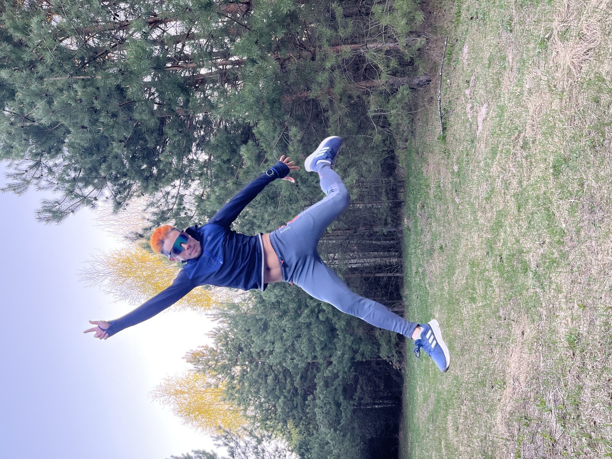 A man with orange hair and blue sunglasses is in mid-air in a grassy area with trees. He is wearing a blue jacket, gray pants, and blue sneakers with white stripes. His arms are outstretched, and his body is angled. The location appears to be a park or grassy area, based on the presence of trees and grass. Based on the prompt, the location is in the city of Chaikovsky, Russia.