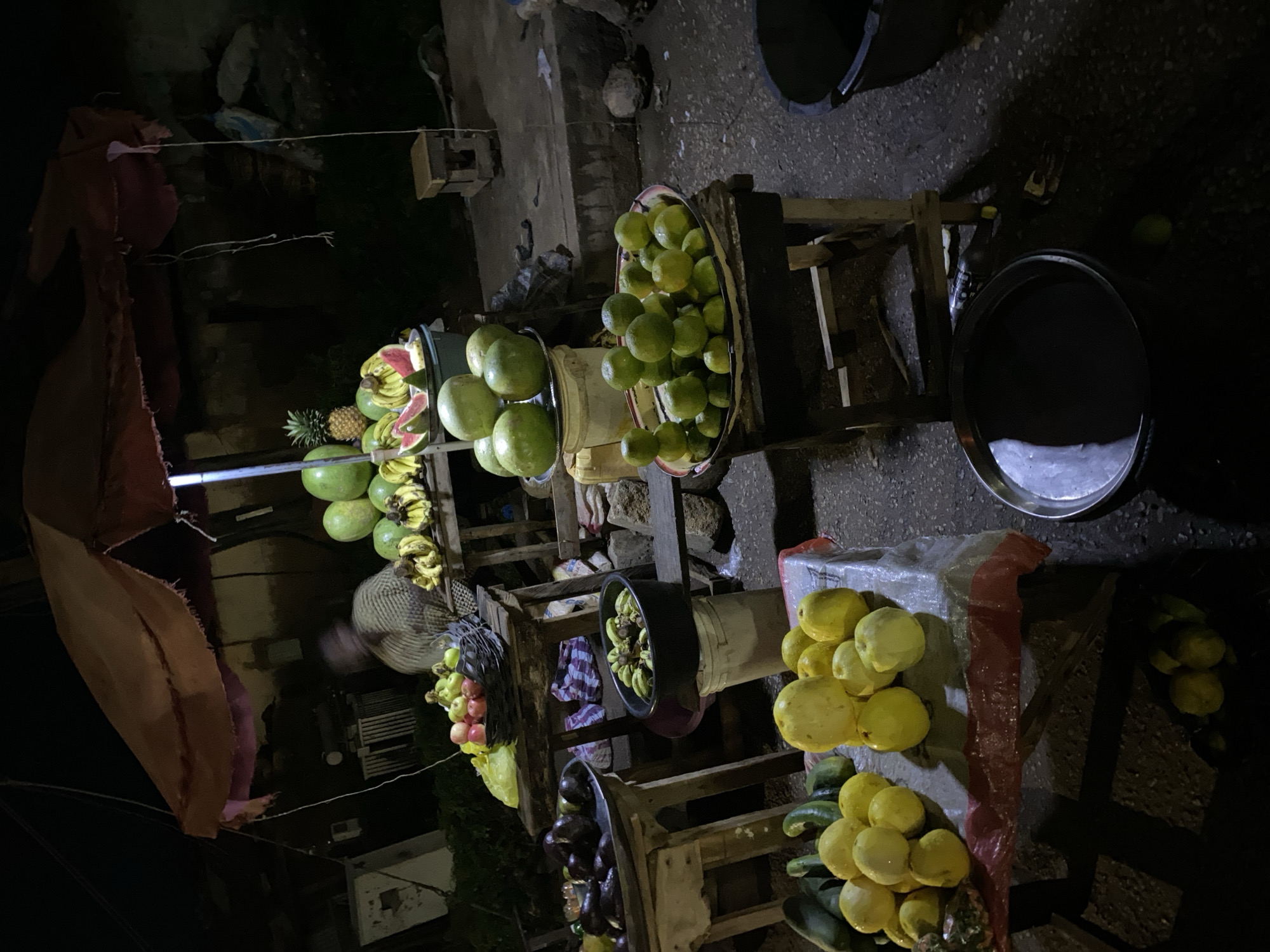The image depicts an outdoor market stall at night in Bukuru, Nigeria. A vendor, whose face is blurred, is seated behind a display of various fruits and vegetables. The stall is covered by a tattered, brown awning supported by poles. On the stall, there are piles of watermelons, pineapples, bananas, oranges, guavas, and eggplants. Some fruits are displayed on wooden tables, while others are in buckets or on metal trays. In the foreground, there is a large metal basin filled with dark liquid. The ground is paved with concrete, and the scene is illuminated by artificial light, creating strong shadows.