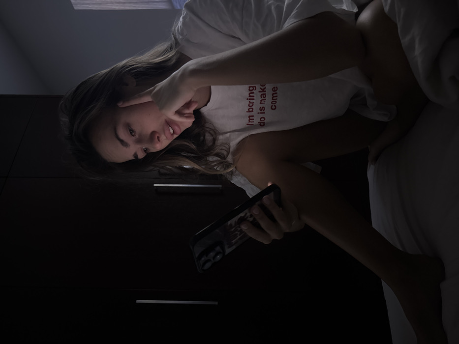 A woman is reclining, holding a phone, with her face angled towards the camera. She is wearing a white t-shirt with red text that reads "I'm boring do is make come". Her left hand touches her face near her cheek. The room appears dimly lit, with a wardrobe visible in the background and bedding nearby. The photograph is rotated, giving a tilted perspective.