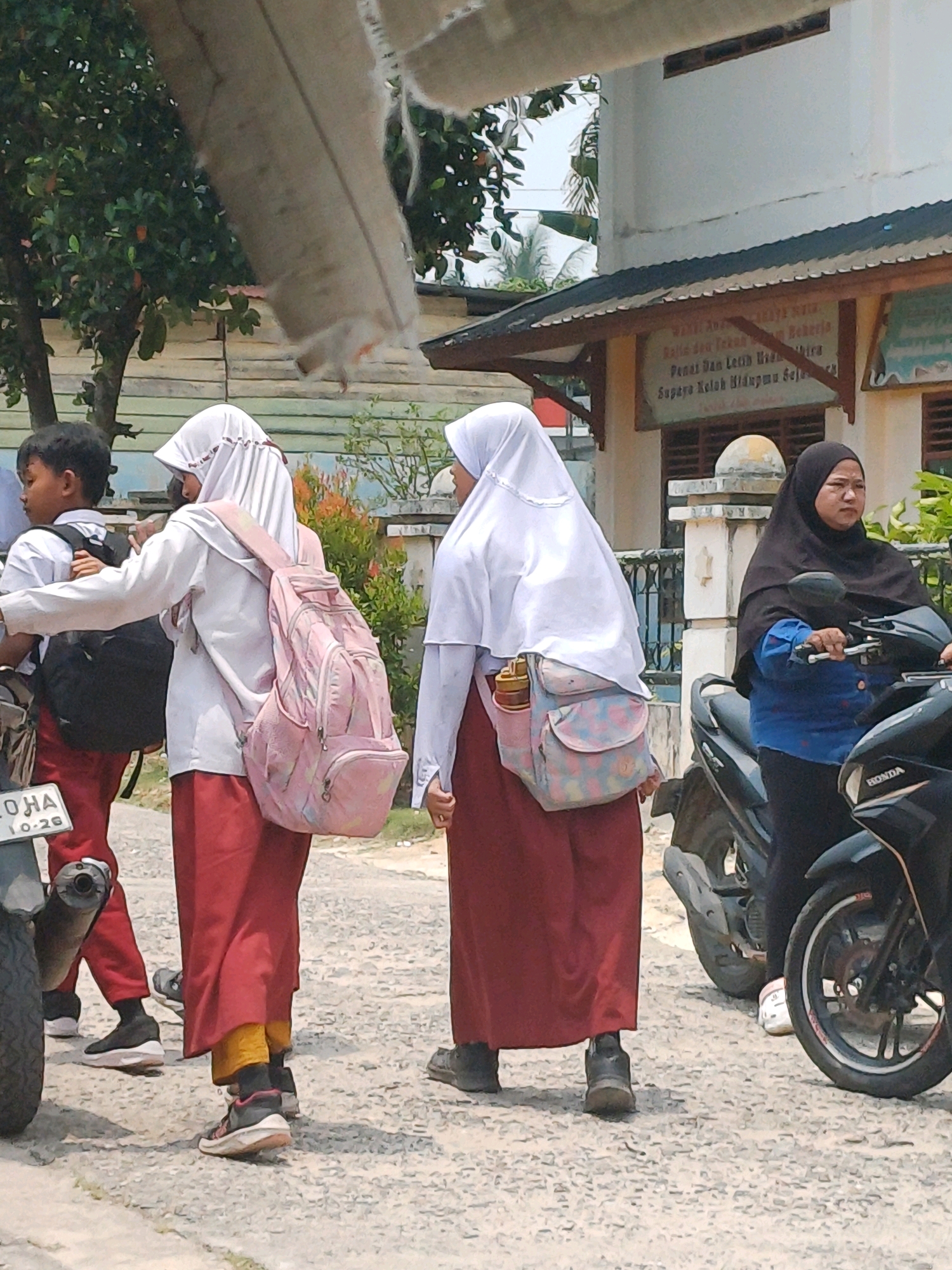 The image depicts a street scene in Dumai City, Indonesia, likely near a school. In the foreground, three individuals are visible. Two are young people, appearing to be students, wearing white shirts and red skirts, with one also wearing a white hijab. They carry backpacks, one of which is pink. The third person is an adult woman wearing a black hijab and a blue shirt, riding a black motorcycle. The individuals are on a gravel road with some vegetation and buildings in the background. The overall atmosphere suggests an everyday moment on a street in the city.