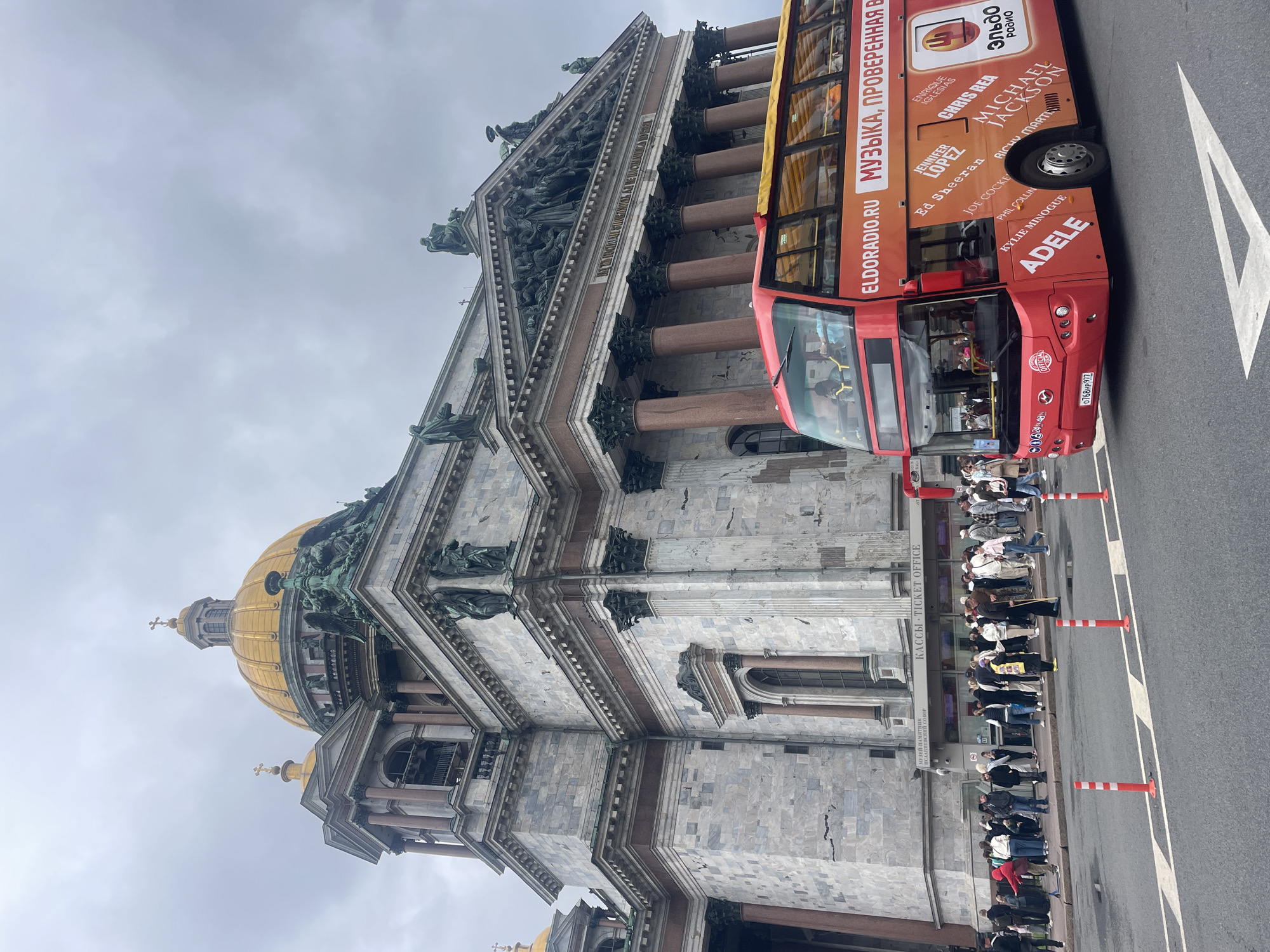 This image captures a view of St. Isaac's Cathedral in Saint Petersburg, Russia, on an overcast day. In the foreground, a red double-decker tourist bus with various artist names and "ELDORADIO.RU" and "МУЗЫКА, ПРОВЕРЕННАЯ ВРЕМЕНЕМ" (Music Verified by Time) printed on its side, is parked on the street. The bus is oriented diagonally, facing away from the viewer. The cathedral's imposing facade, made of marble, features large columns, intricate sculptures, and a prominent golden dome. A crowd of people is gathered outside the cathedral's ticket office, indicated by signs in Russian and English. Orange and white traffic cones and a white directional arrow are visible on the asphalt road. The sky is cloudy, creating a subdued lighting condition.