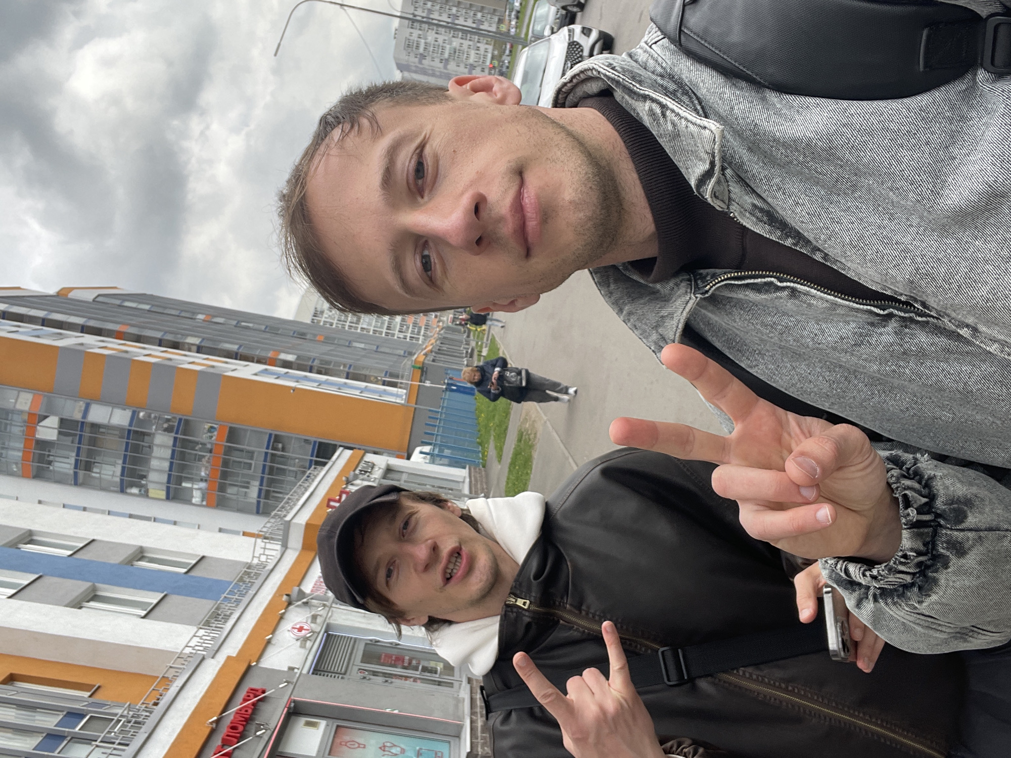 The image shows two men posing for a selfie in front of a building in an urban setting. The man in the foreground wears a grey denim jacket and a black bag. He is holding his fingers in a "peace" sign. The other man, behind and to the left, wears a black jacket and cap and flashes a "rock on" hand gesture. In the background, there is a modern building with orange, white, and blue panels, street lamps, a sidewalk, and a person walking in the distance. The sky is overcast. There are also signs visible on the side of the building.