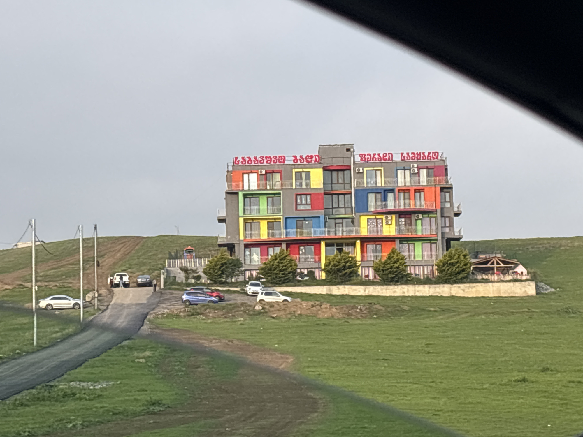 The image shows a five-story building on a grassy hill, seemingly taken from inside a vehicle. The building is a hotel or apartment complex, with colorful facades of red, blue, yellow, green, and orange. There's text in red lettering above the building, presumably in Georgian script. A driveway leads up the hill to a parking area where several cars are parked. There are trees and other low-lying vegetation around the building. The sky is overcast. The road in the foreground appears to be asphalt. The specific location in Tbilisi, Georgia, is not determinable solely from the visual information provided in the image.