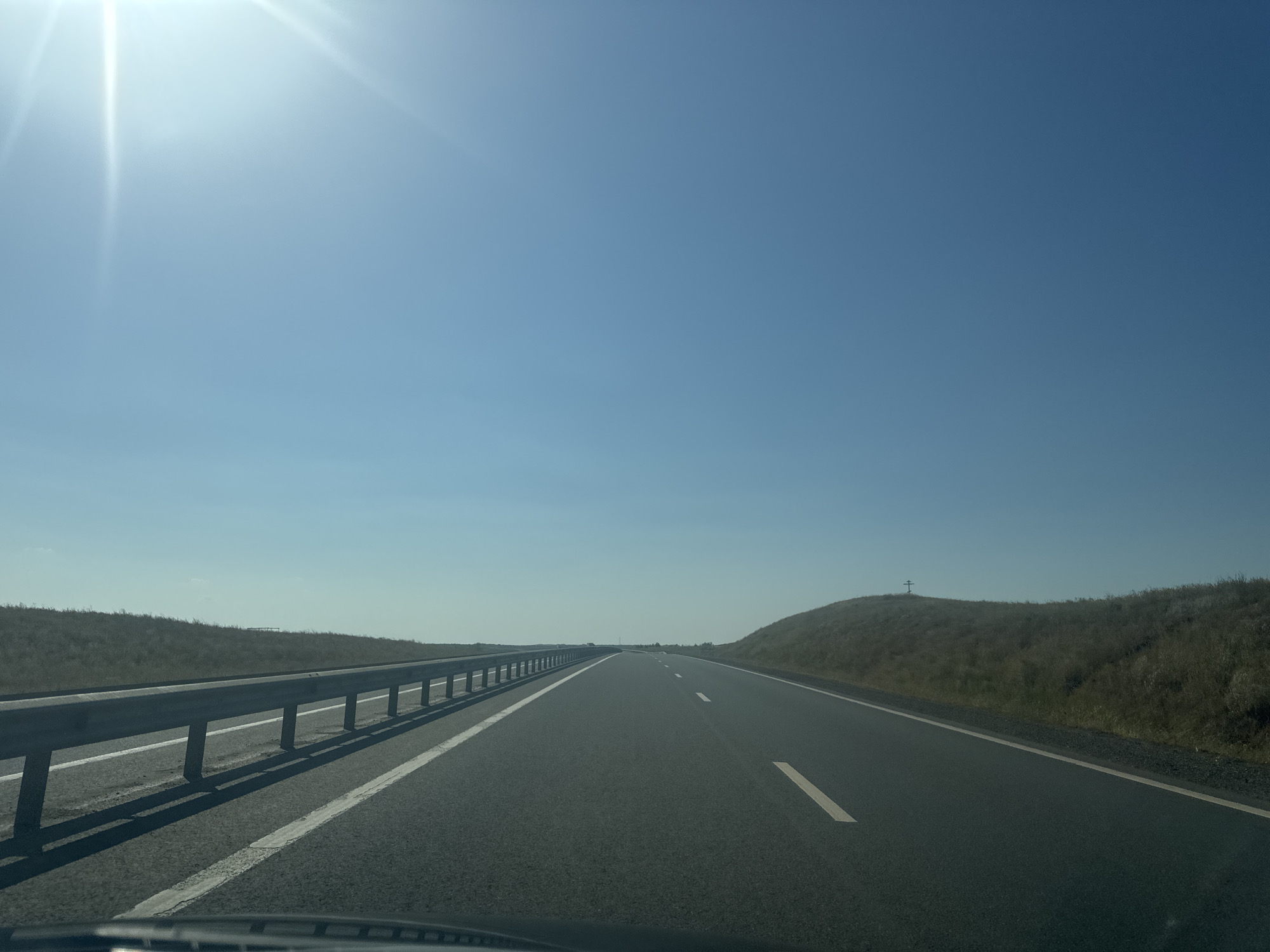 The image depicts a view from the driver's perspective of a highway, likely a road in or near Syrt, Russia, given the context provided, though specific Syrt landmarks are not identifiable from the image alone. The sky is clear and a bright blue, with the sun prominently visible and casting rays in the upper left quadrant. A highway guardrail runs along the left side of the road, separating it from a grassy embankment. The highway itself is asphalt with white lane markings. On the right side, another grassy embankment rises, with a small, dark cross-shaped structure visible atop it. The landscape appears arid or sparsely vegetated, consistent with open terrain. No people or vehicles are directly in focus or prominently featured in the image, besides the implied presence of the photographer's vehicle.