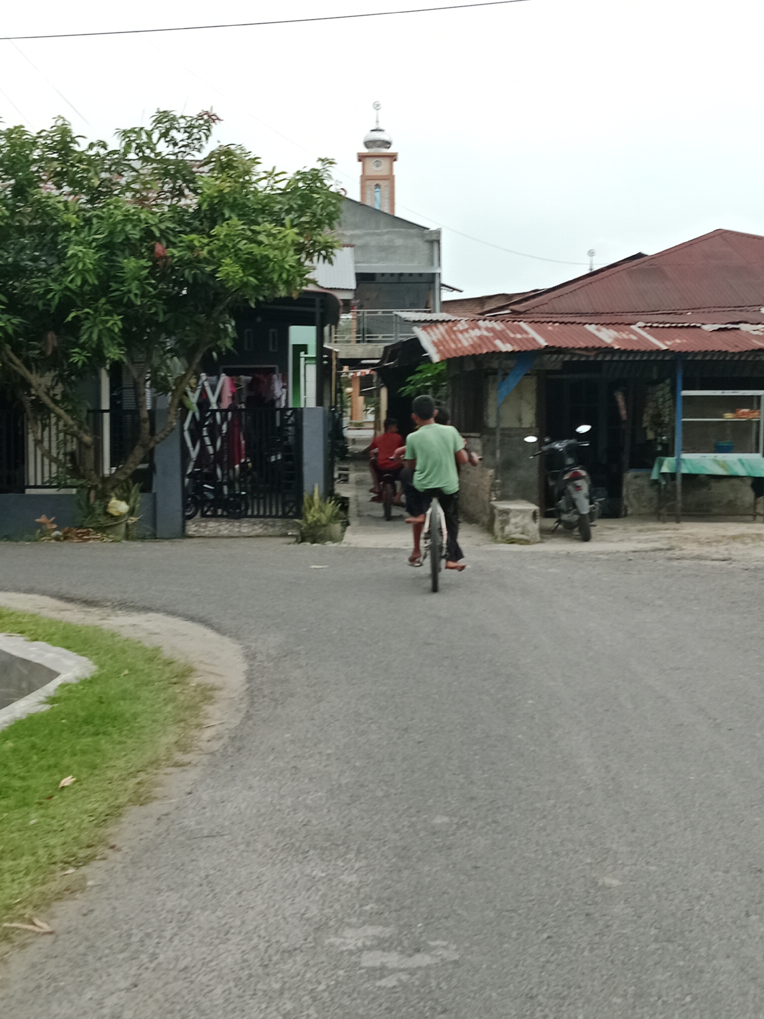 In the City of Binjai, Indonesia, on an overcast day, a young boy with dark hair rides a bicycle down a paved road. He is wearing a light green t-shirt and dark shorts. Behind him, another boy in a red shirt is also on a bicycle. The road is bordered by a drainage ditch and a strip of grass on the left, and houses and businesses on the right. One house has a grey facade and a black gate with laundry hanging behind it. Another building to the right has a rusty corrugated metal roof and appears to be a shop with a blue table and window display. In the background, a mosque with a tall, light-colored minaret can be seen. A motorcycle is parked in front of one of the buildings. The overall scene depicts a residential street in a tropical climate.