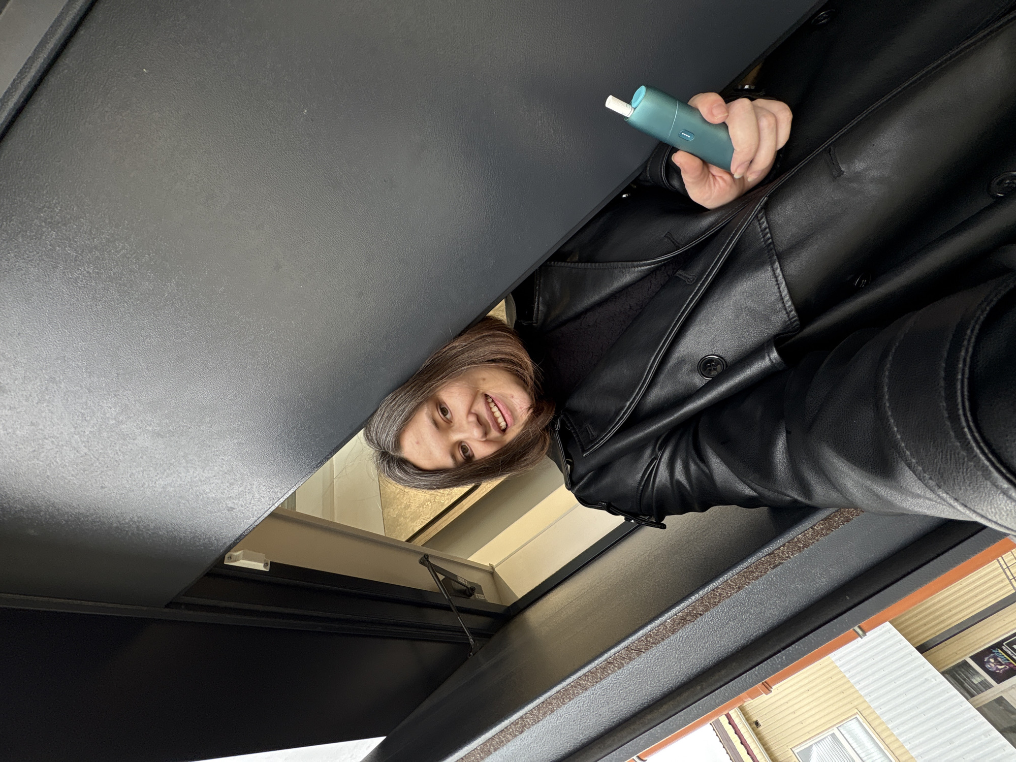 A woman is pictured from an upward angle, likely a selfie, holding a teal-colored electronic cigarette. She is wearing a black leather coat. Part of the interior of a structure, possibly a sunroof, is visible. Beyond the roof is a building with light yellow siding and white trim, suggesting the location is Chaikovsky, Russia.