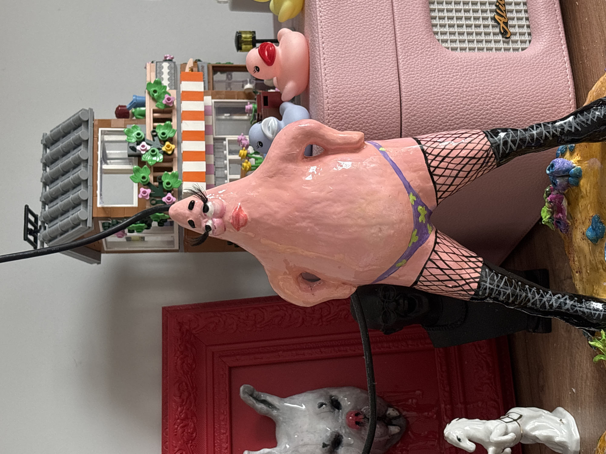 The image shows a collection of objects arranged on a surface. From left to right, there's a building made of what looks like Lego bricks, decorated with plastic plants and flowers. Next to it is a pink, rectangular, structured bag with a white-backed logo with golden letters. In front of the bag is a bizarre pink figurine wearing black fishnet stockings and boots and a purple swimsuit. Beneath the figurine, there's a red ornate picture frame. Inside the frame, there's a white ceramic dog head attached to the back of the frame with another smaller white ceramic dog on the bottom right corner. The objects are placed against a white wall.
It is impossible to determine the location in the city of Krasnoyarsk, Russia, from this image.